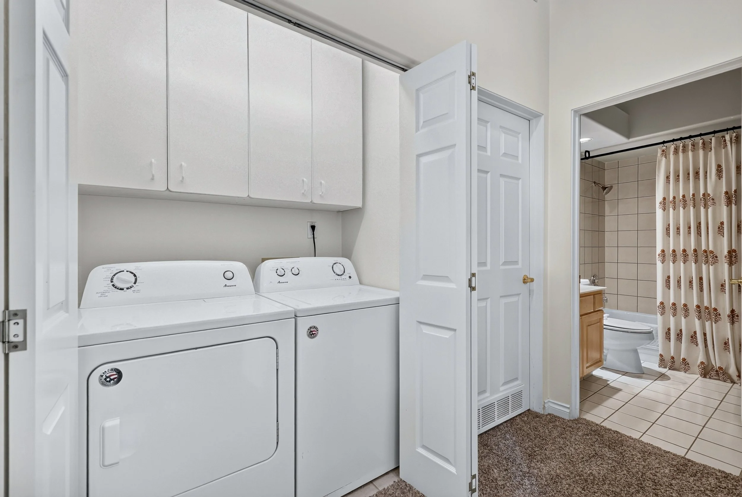 A laundry room with a white washer and dryer, upper cabinets, and open doors leading to a bathroom with a shower, shower curtain, toilet, and bathroom vanity.