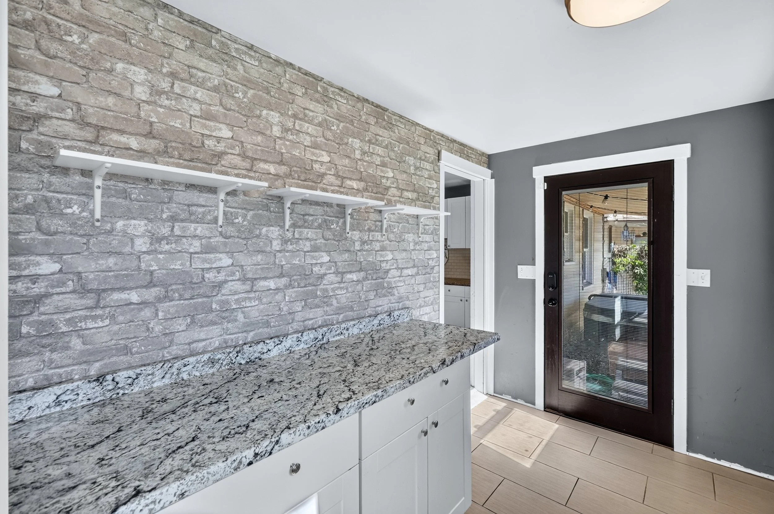 Kitchen with gray brick wall, granite countertop, white cabinets, and a glass door leading outside.