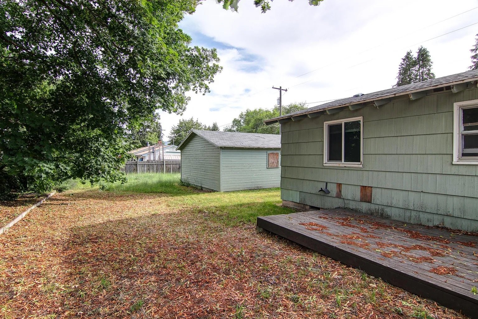 Backyard scene showing a weathered green house with peeling paint, a deck with fallen leaves, grass, a tree with dense green leaves, a small shed, and a wooden fence in the background under partly cloudy sky.