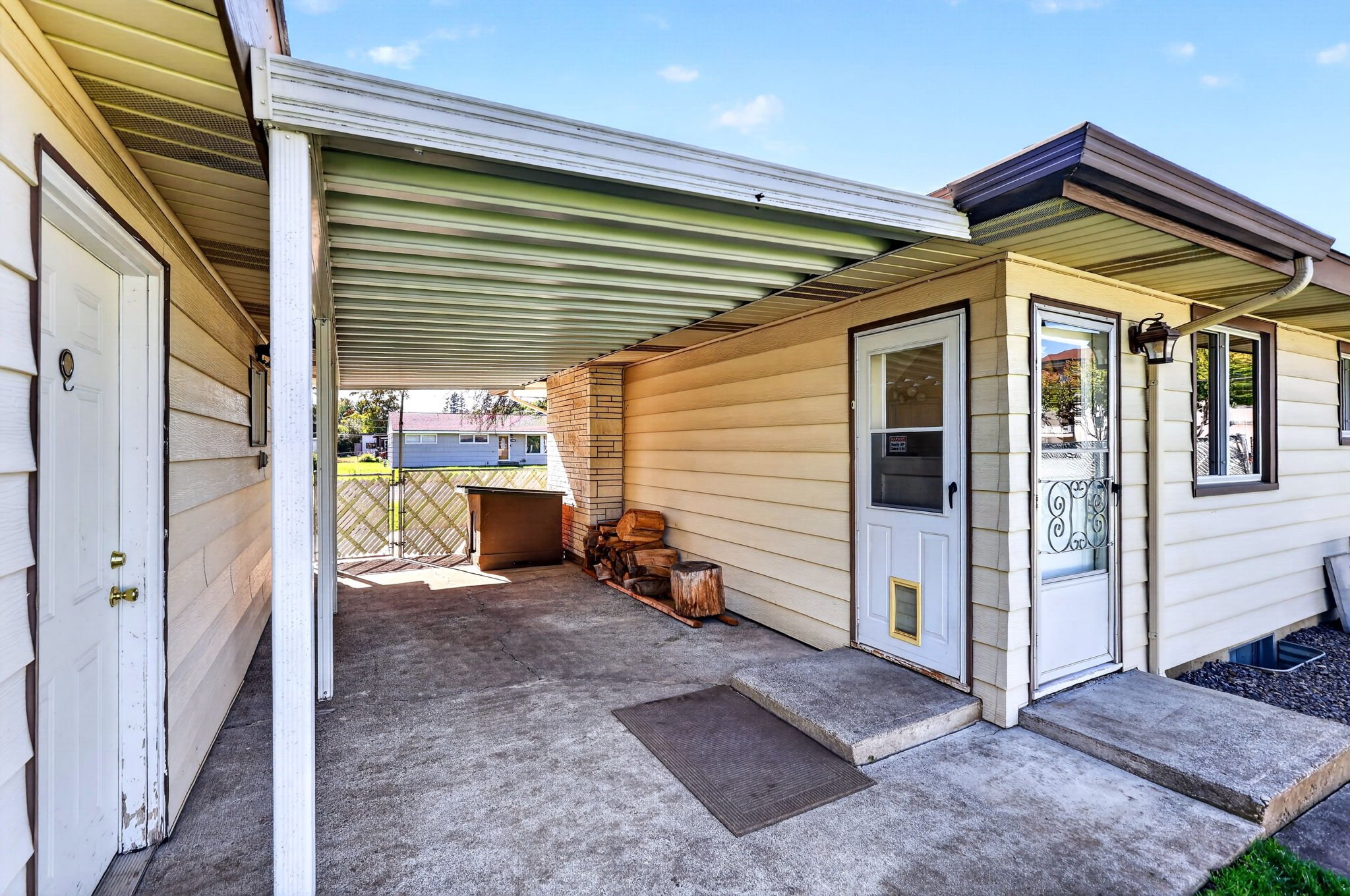 Back patio area of a house with yellow siding, a sliding glass door, a small window, and a wood storage pile, covered by a metal roof. There is a light fixture on the wall and a white door with a gold knob.