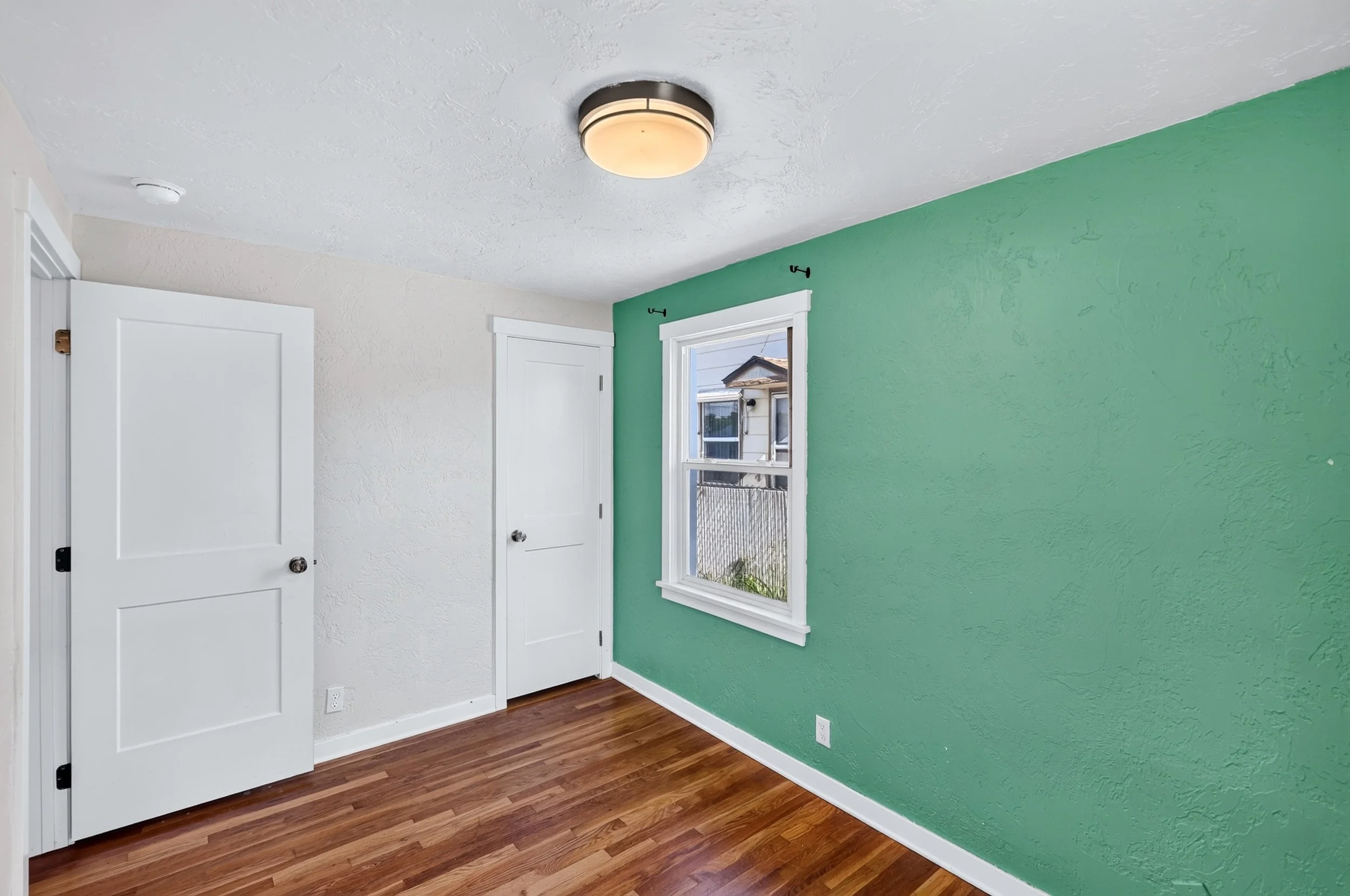 Room with green accent wall, white doors, and a window showing a house outside. The floor is wood, and there is a ceiling light fixture.