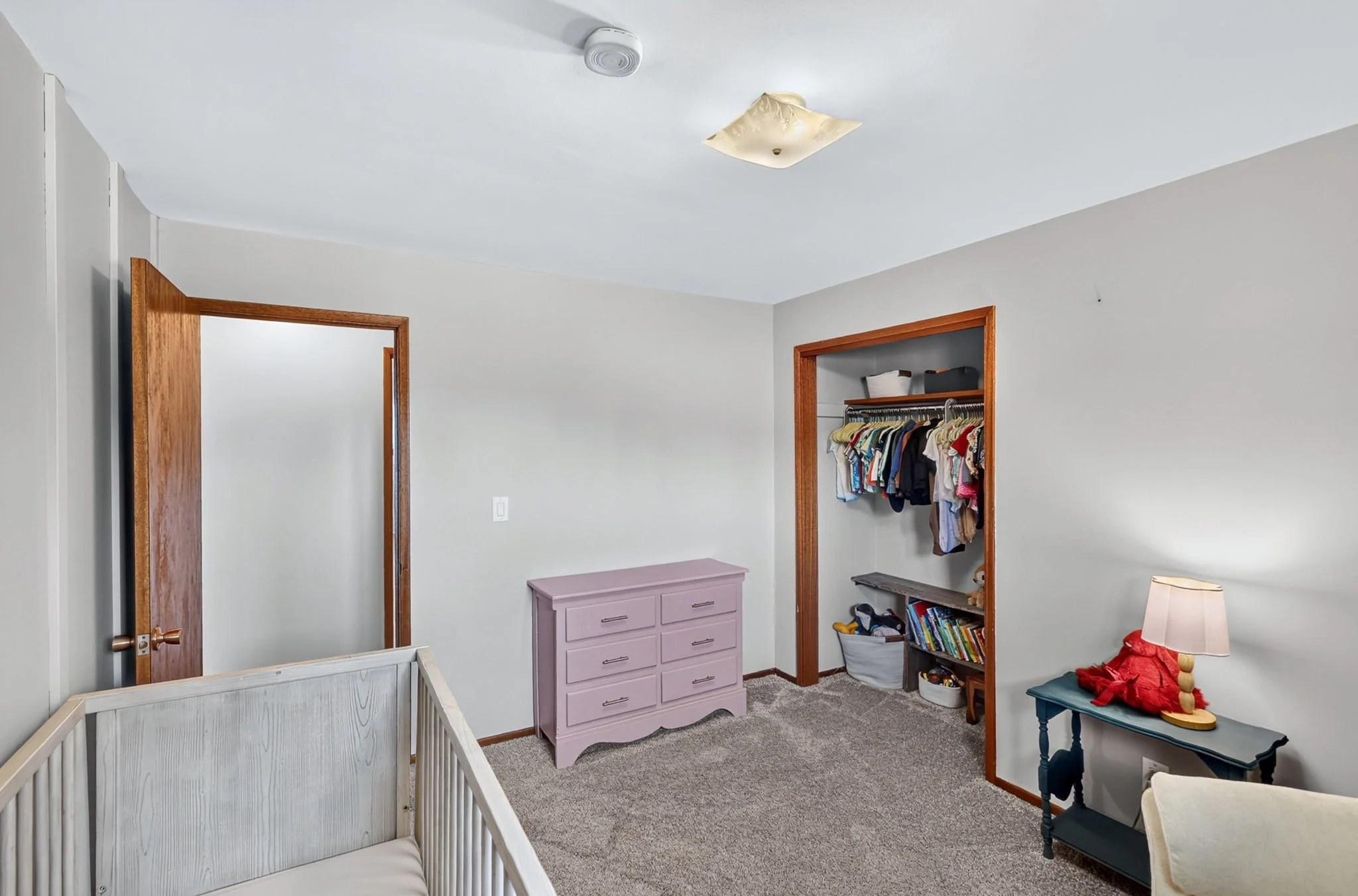 A bedroom with a white crib, a purple dresser, a small black bookshelf, and an open closet with clothes hanging. The room has white walls, a tan carpet, and two wooden doorways.
