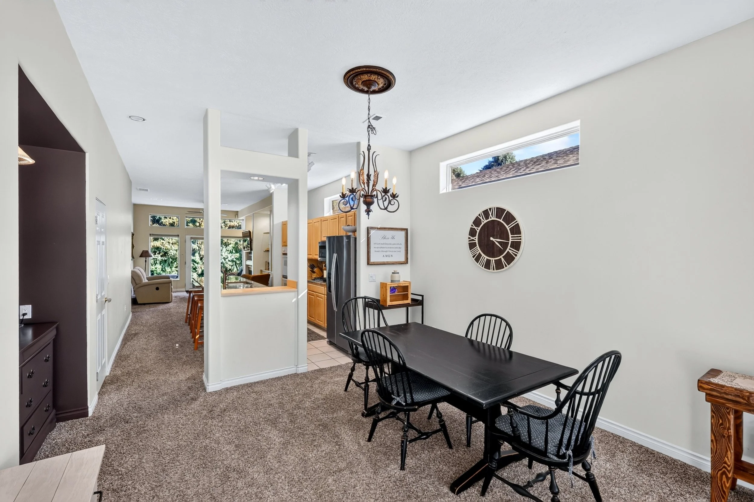 Open-concept living room, kitchen, and dining area with a black dining table and chairs, beige carpet, and large windows letting in natural light.
