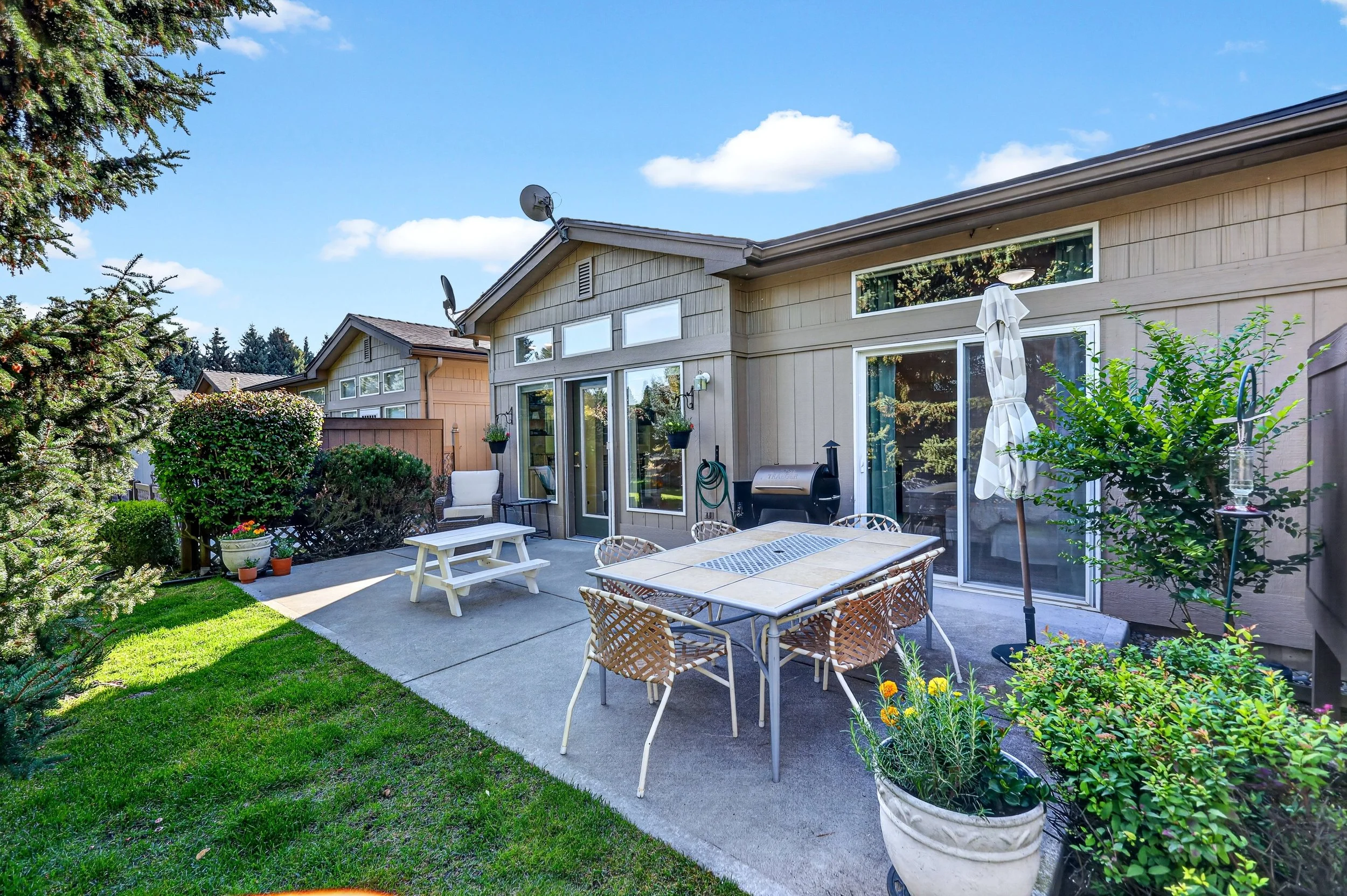 A backyard patio with a dining table, chairs, an umbrella, potted plants, lush green grass, and a view of neighboring houses under a partly cloudy sky.
