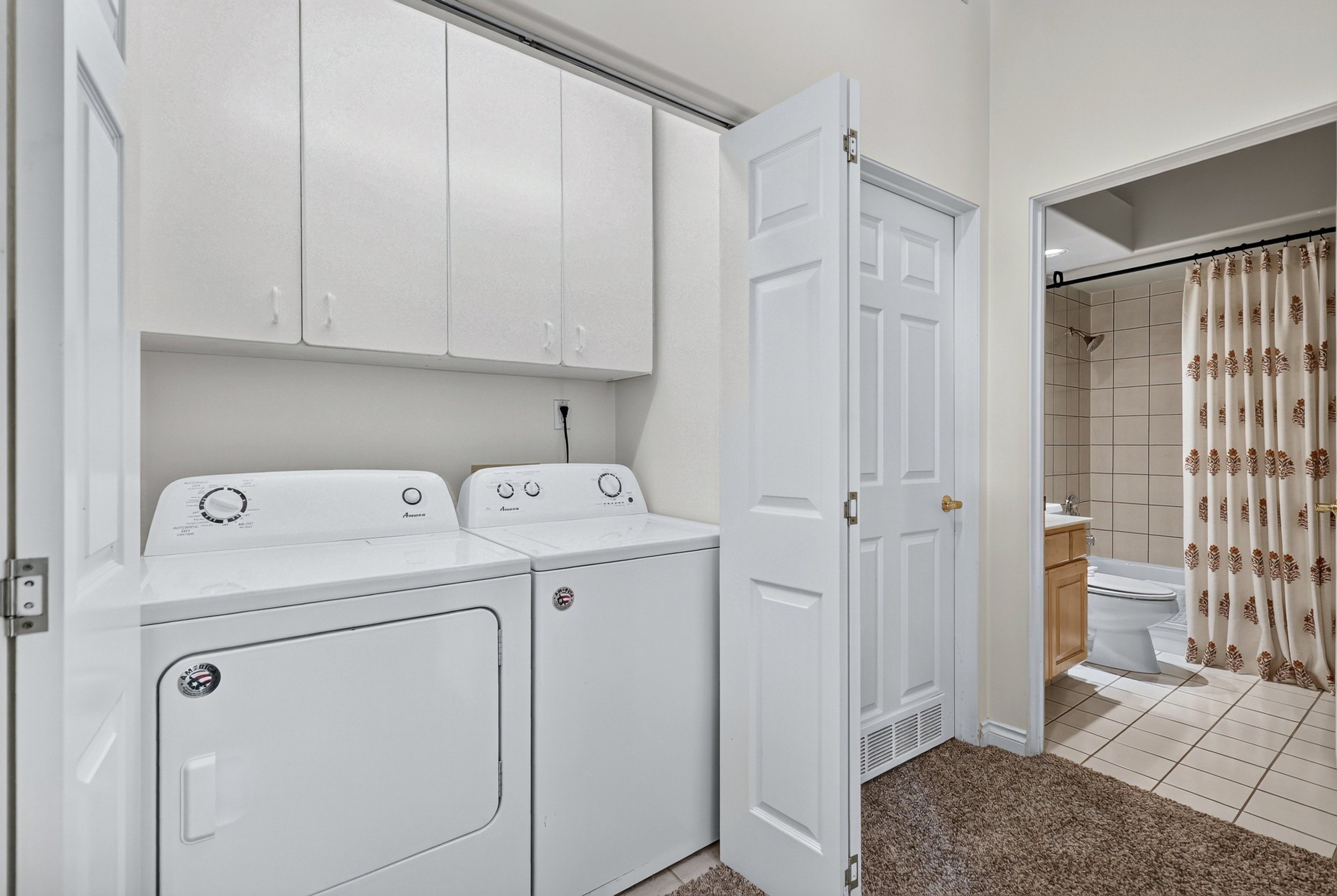 A laundry area with a white washing machine and dryer, white cabinets above, with a bathroom visible through an open door in the background, featuring a bathtub with a shower curtain and tiled walls.