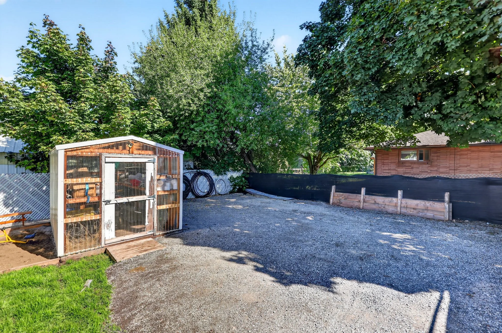 Backyard with trees, a shed, a gravel path, and a wooden fence, under a clear blue sky.