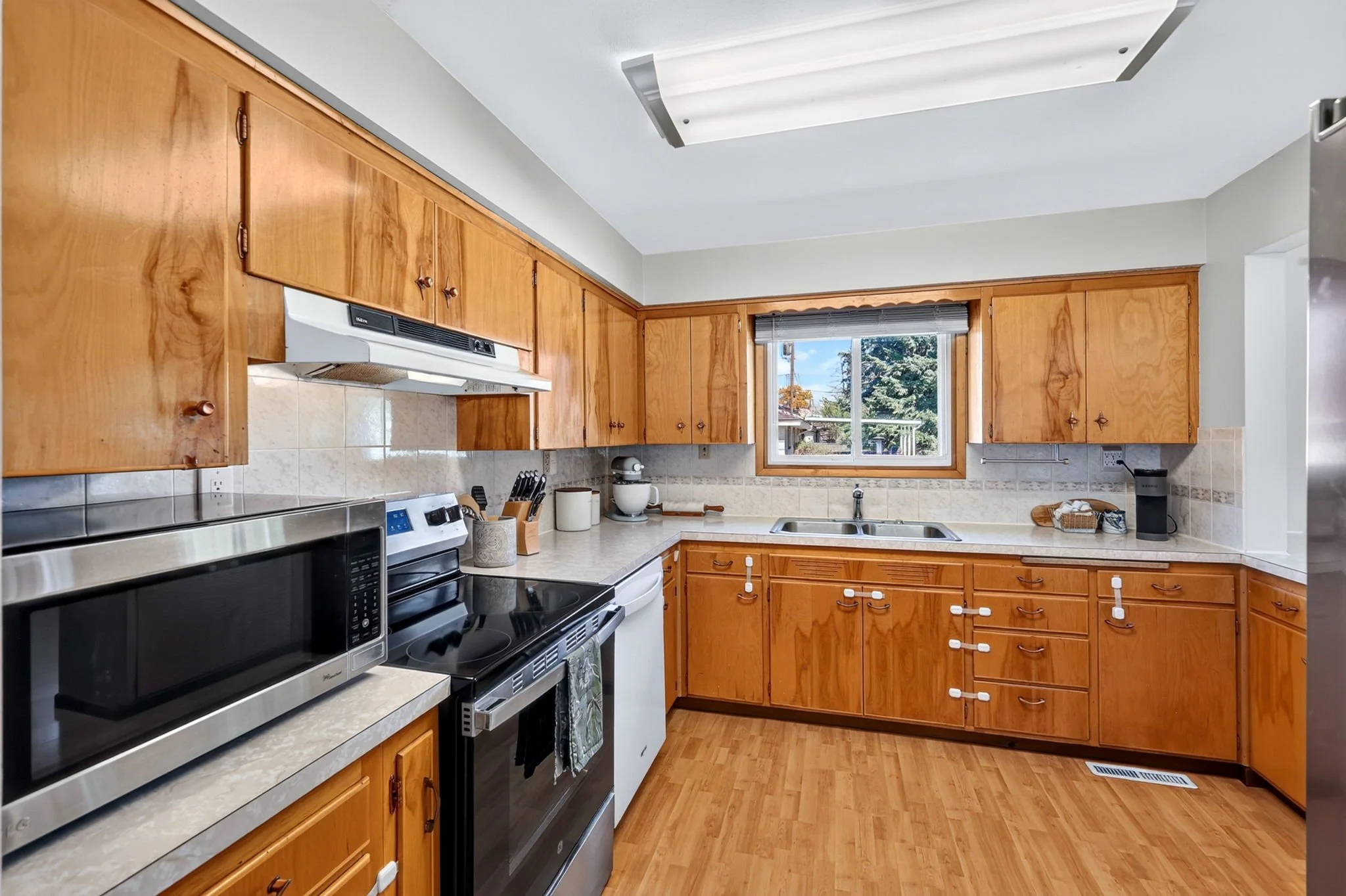 A kitchen with wooden cabinets, a window above the sink showing trees outside, a stove with an oven, microwave, and various kitchen items on the countertop.