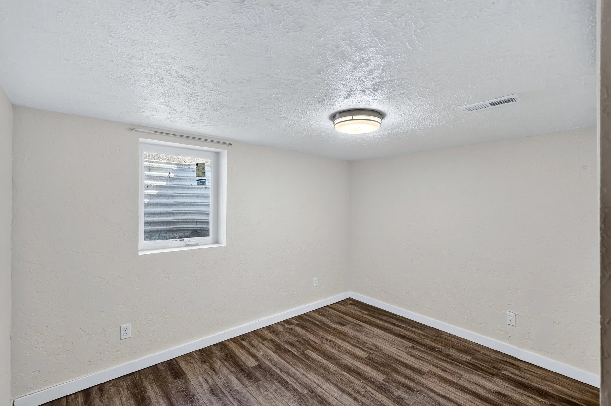 Empty room with beige textured walls, a small window, a ceiling light fixture, and hardwood flooring.