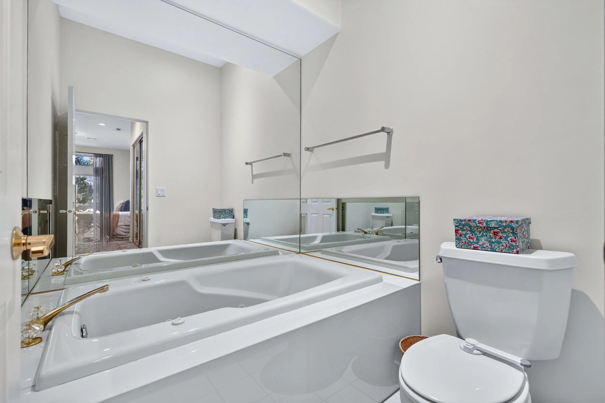 A bathroom with a large mirror, white bathtub with gold fixtures, a toilet with a floral tissue box on top, and a view into a bedroom through a doorway.