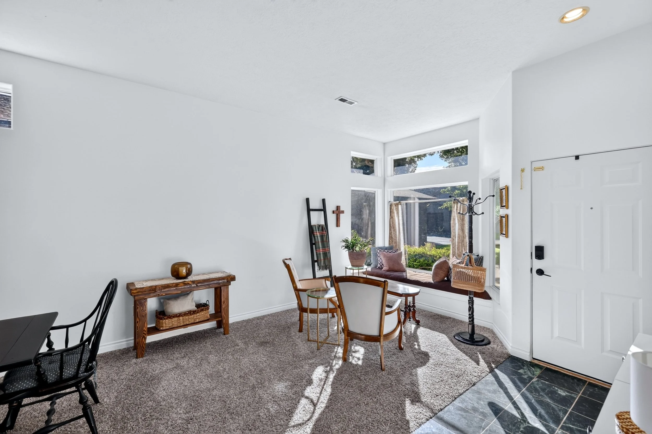 Bright living room with large windows, white walls, and a mix of furniture including chairs, a wooden table, and a window seat with cushions. Light and shadow create a cozy atmosphere.