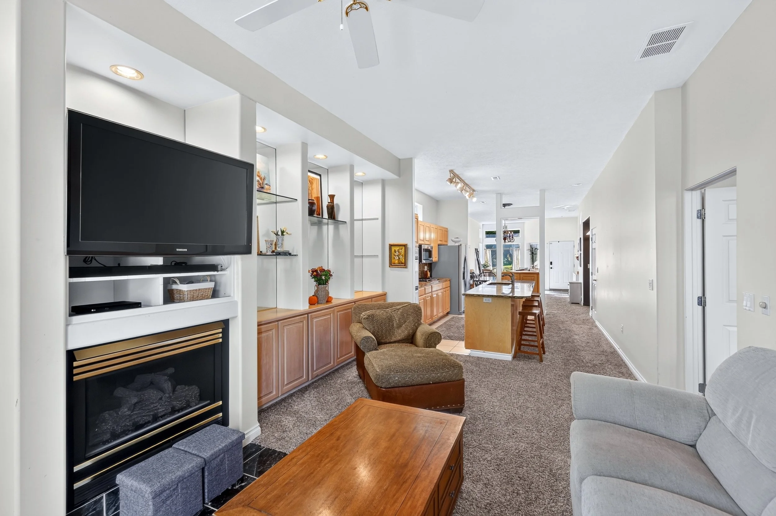 Living room with a fireplace, flat-screen TV, armchair, wooden coffee table, and a sofa. Open kitchen and dining area in the background with wooden cabinets, a refrigerator, and a window with a view outside.