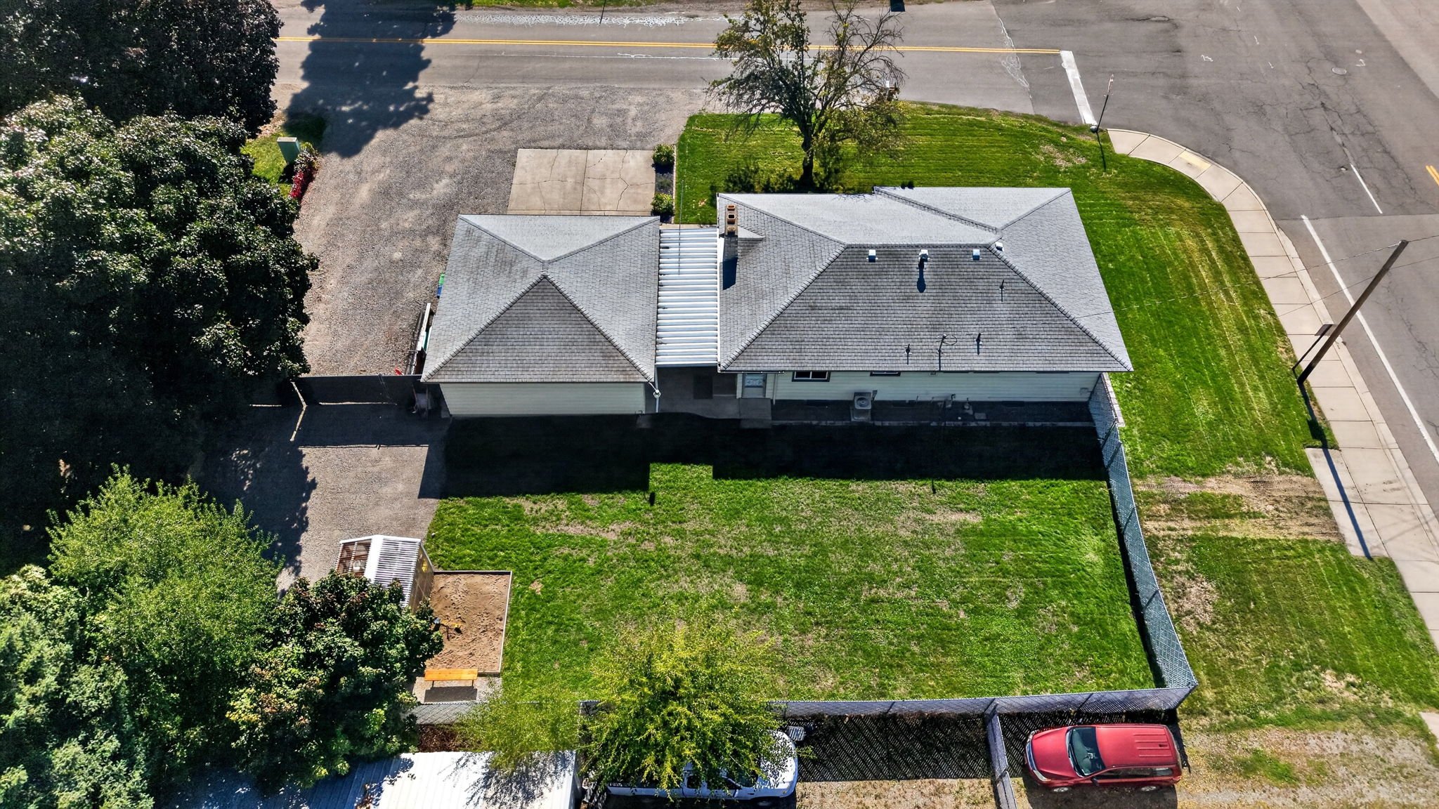 An aerial view of a house with a gray shingle roof, green lawn, trees, a driveway, and surrounding streets.