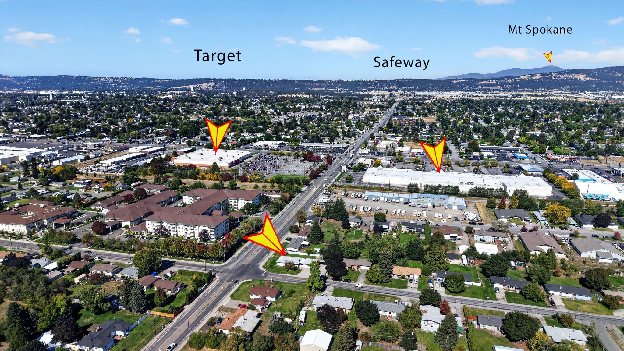 Aerial view of a city with suburban houses, commercial buildings, and a main road, with mountains in the background. The image has labels for Target, Safeway, Mt Spokane, and a direction marker.