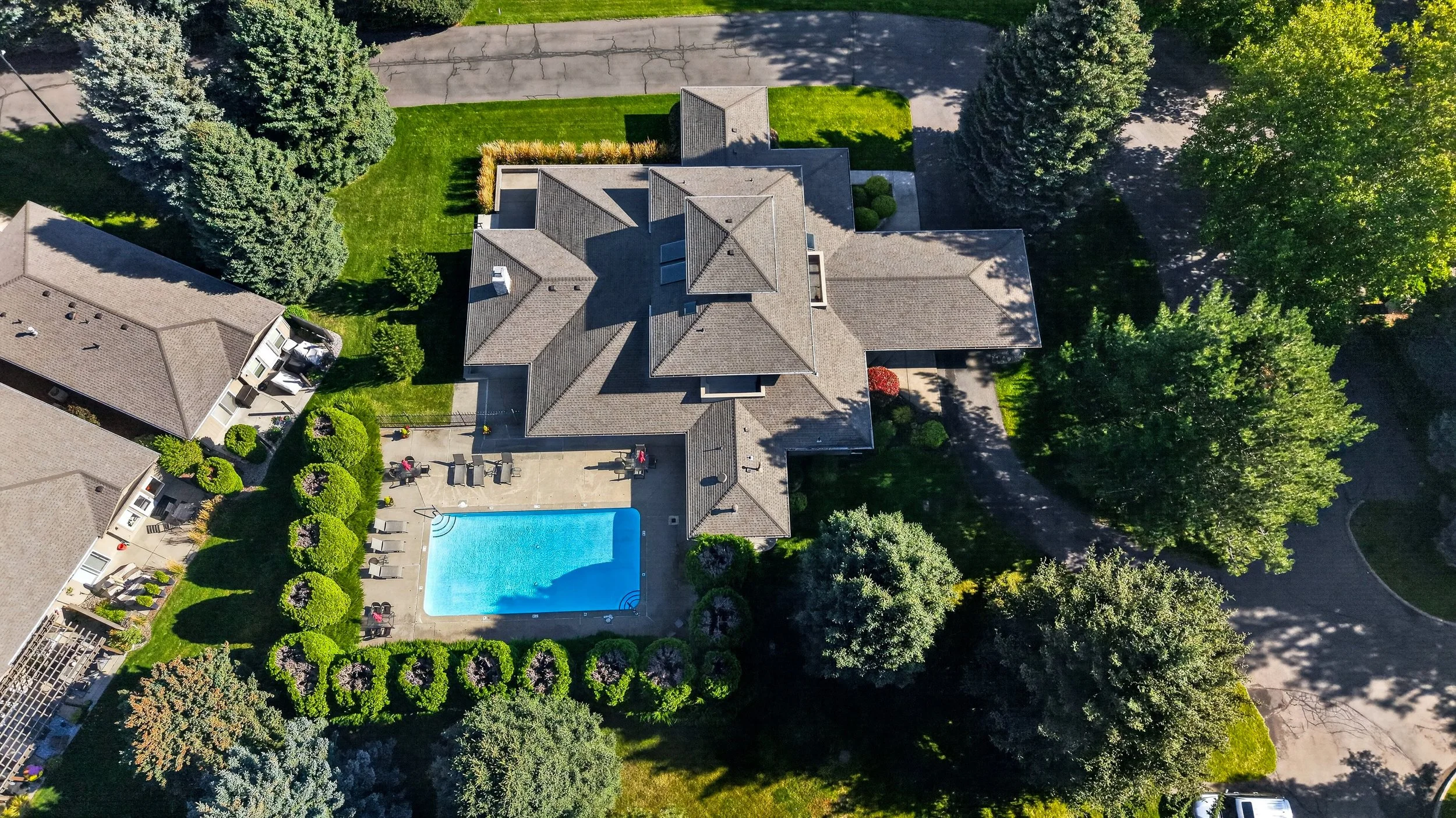 An aerial view of a residential house with a backyard, swimming pool, surrounding trees, a parking lot, and neighboring houses.