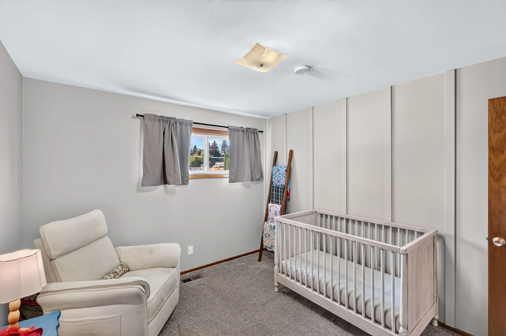A room with a white crib, a white armchair, and a window with gray curtains. There is a wooden folding ladder with clothes, and a small table with toys. The ceiling has a light fixture and a smoke detector.
