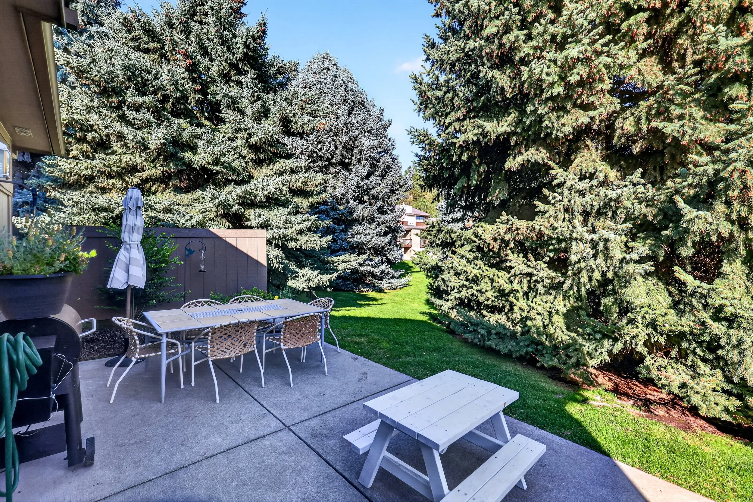 A backyard patio with a white picnic table and benches, a round table with chairs, a closed striped umbrella, lush green grass, and tall pine trees in the background under a clear blue sky.