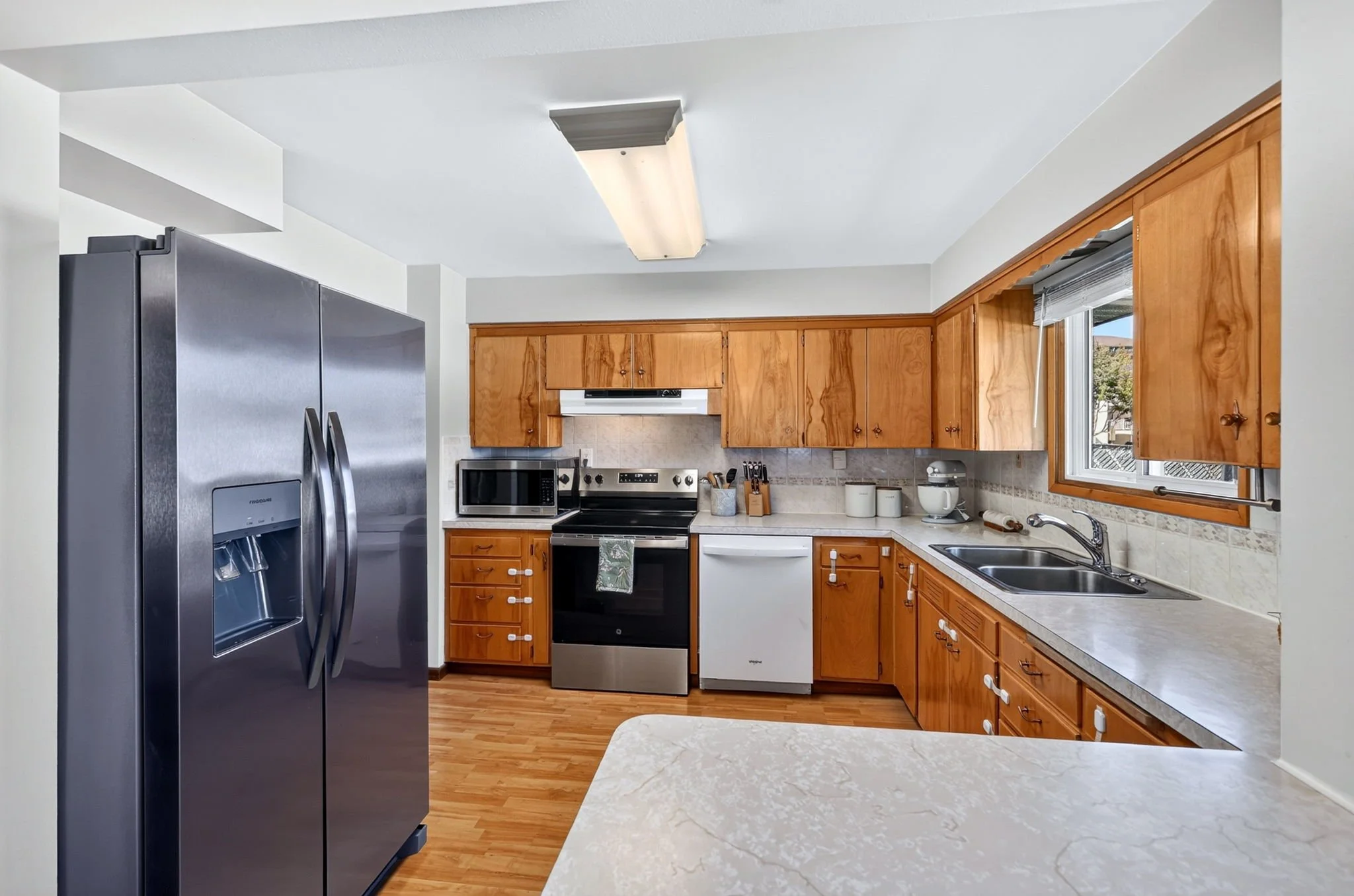 Kitchen with stainless steel refrigerator, wooden cabinets, microwave oven, stove, and double sink next to window with a view outside.