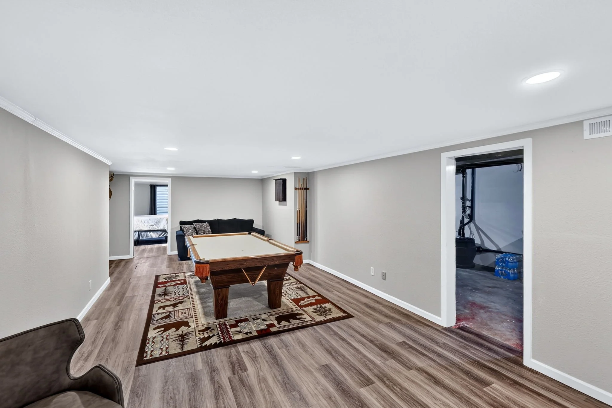 A spacious basement living area with light-colored walls and wood flooring. There is a pool table in the center on a decorative area rug. In the background, it has a sitting area with a dark sofa, a building with large windows, and a doorway to anoth