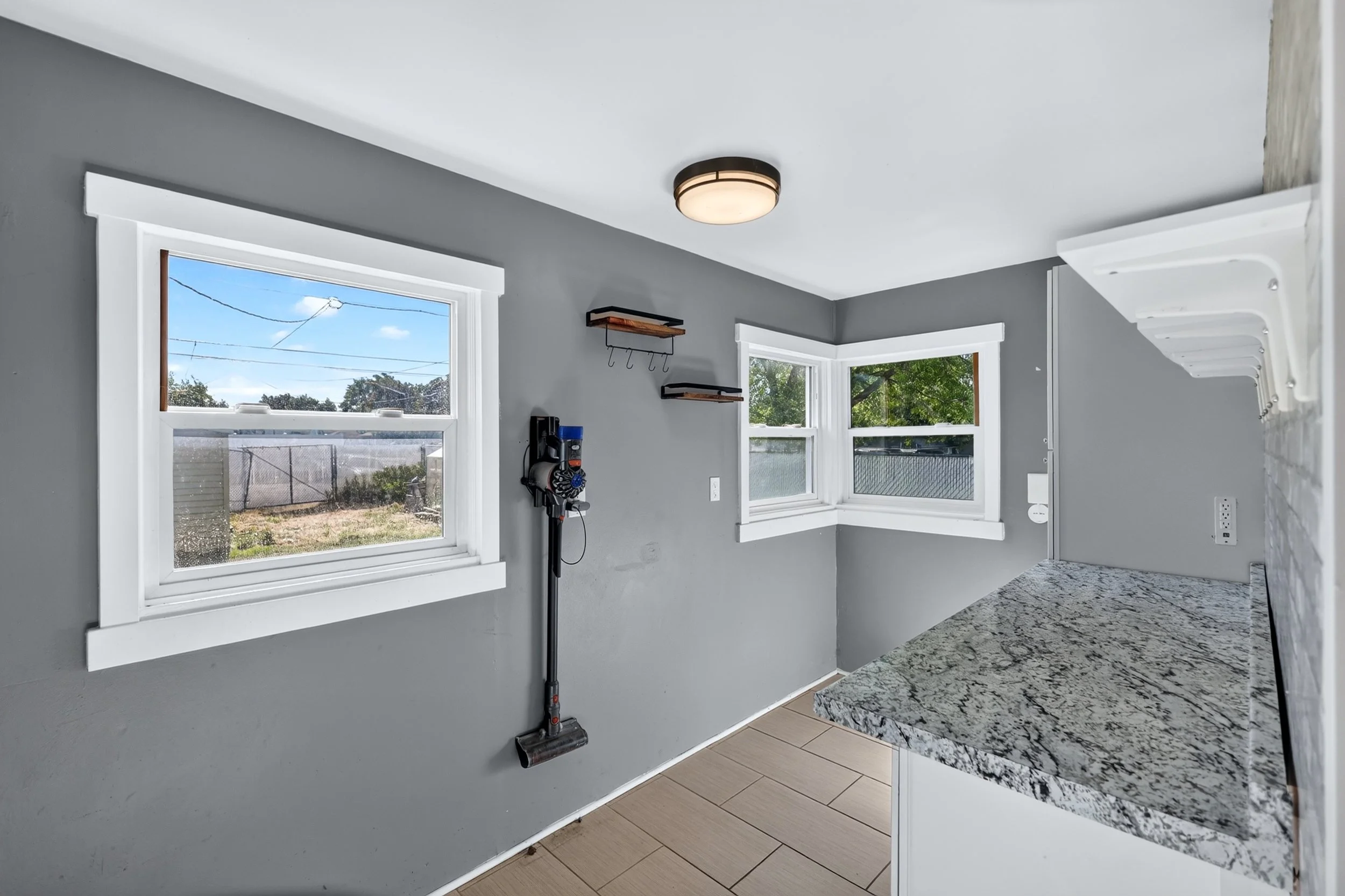 Clean, modern kitchen with gray walls, white trim, granite countertop, and large windows showing outside greenery and blue sky.