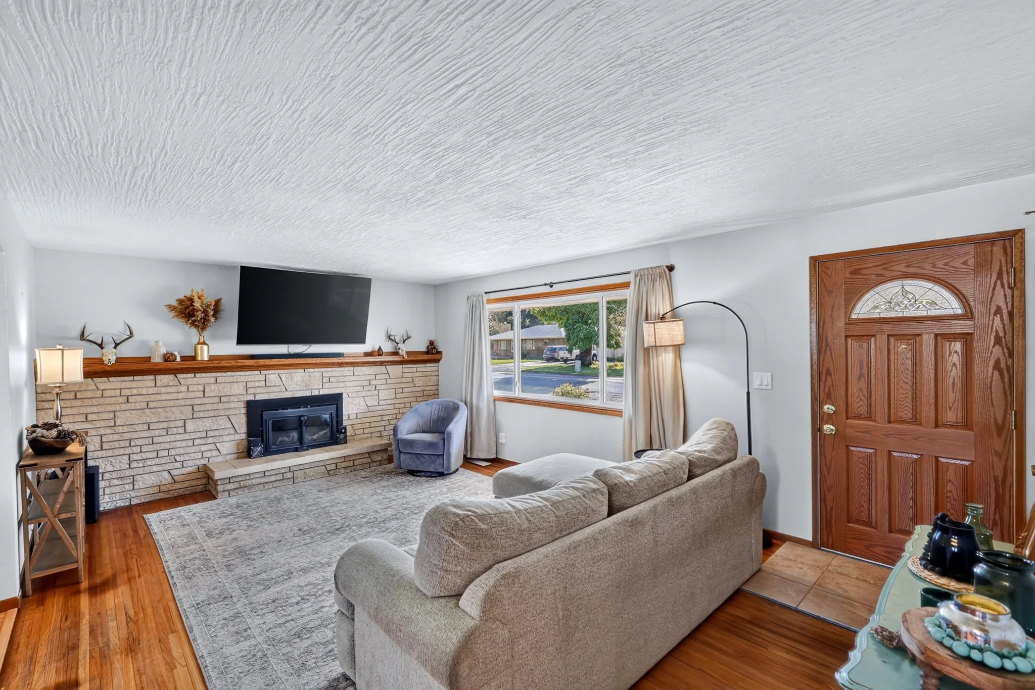 Living room with brick fireplace, wall-mounted TV, beige couches, armchair, window with curtains, hardwood floor, and decor like antlers, vases, and lamps.