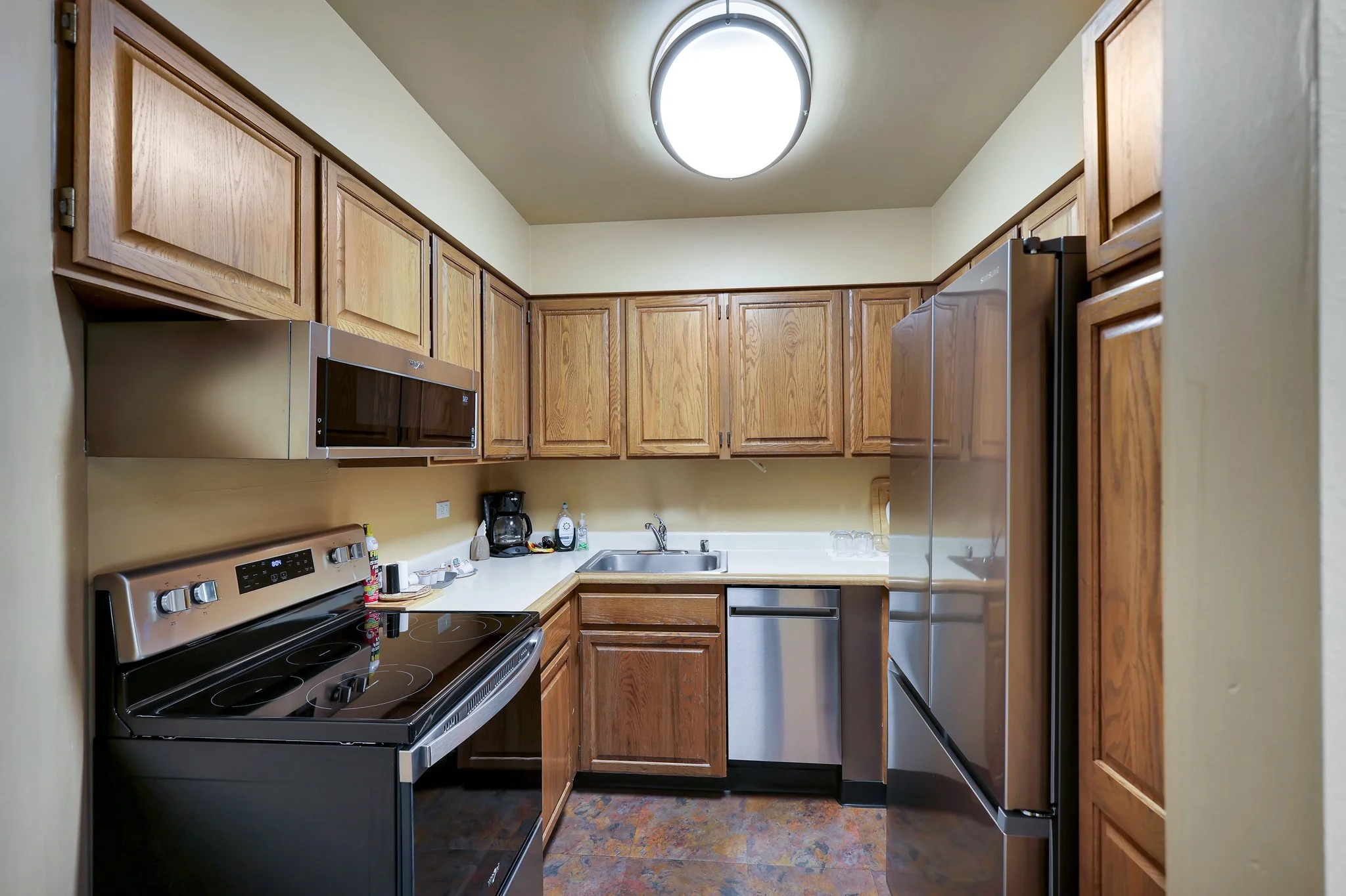 A small kitchen with wooden cabinets, a gray microwave, a stove, sink, coffee maker, and refrigerator.