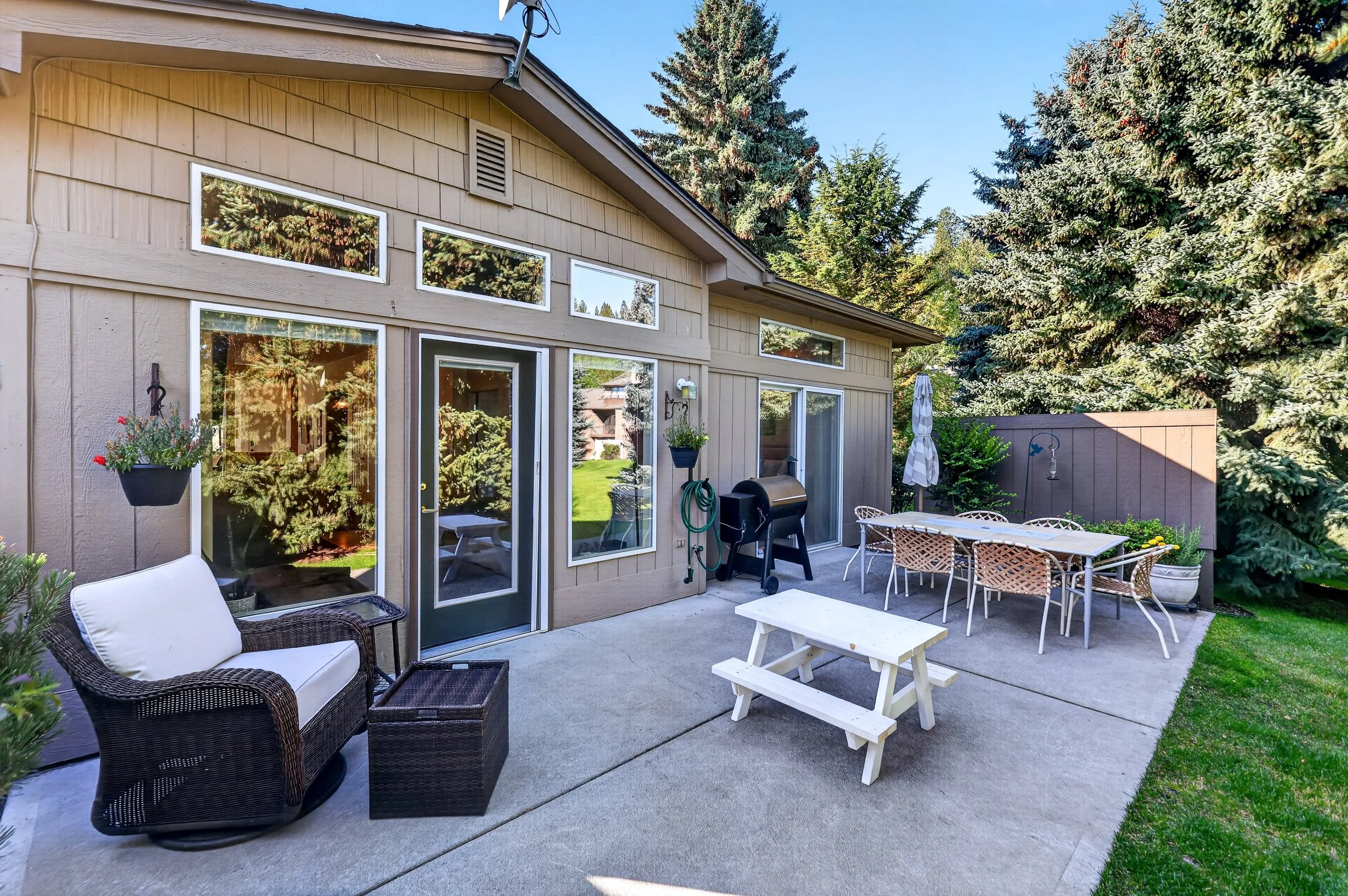 A backyard patio with outdoor furniture including a wicker sofa with cushions, a white picnic table, wicker chairs, and an umbrella. There are potted plants and garden accessories, with large trees providing a lush background.
