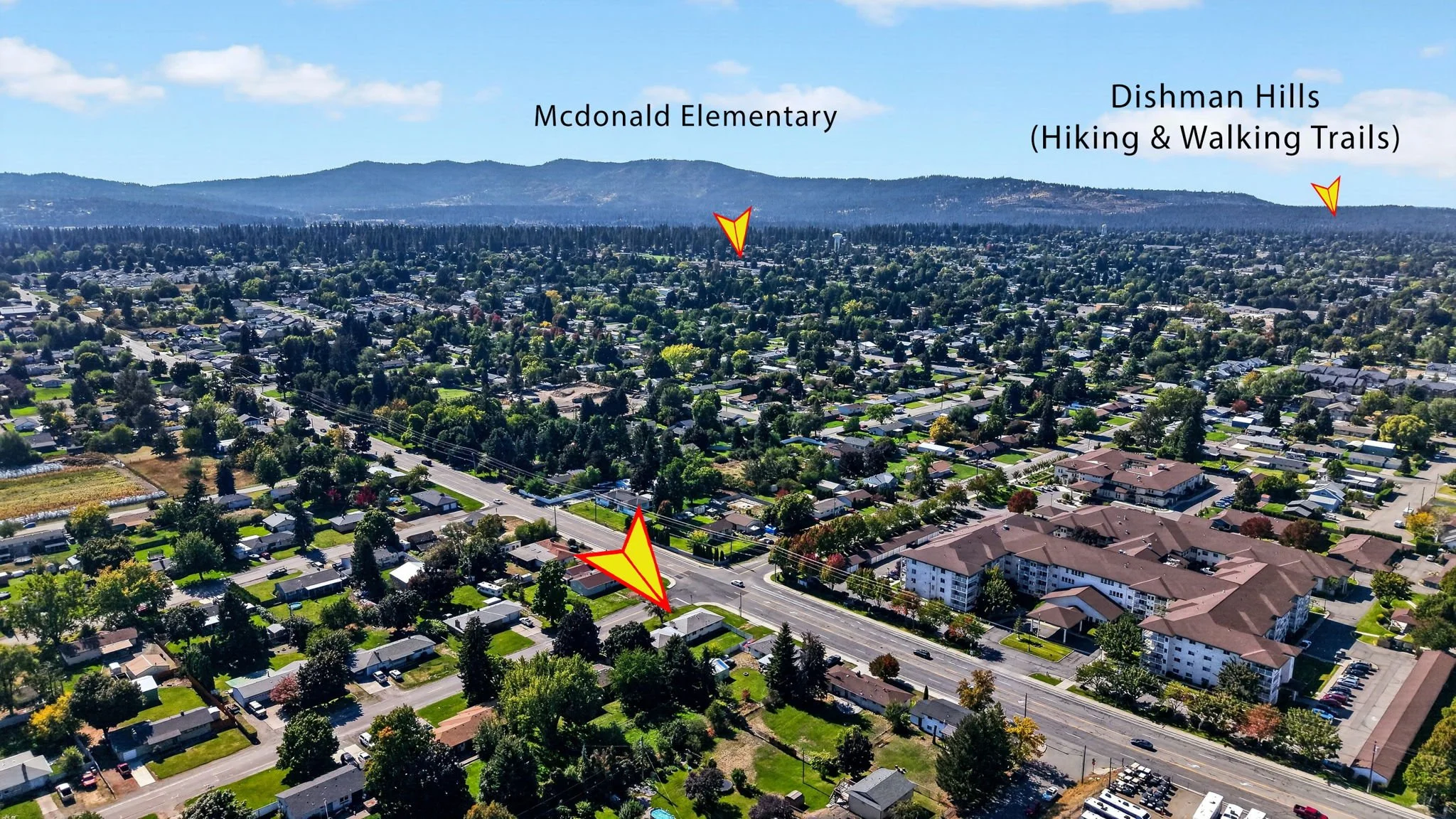 Aerial view of a suburban neighborhood with houses, trees, and a busy street with cars. Labels point out Mcdonald Elementary, Dishman Hills hiking and walking trails, and a road with traffic.