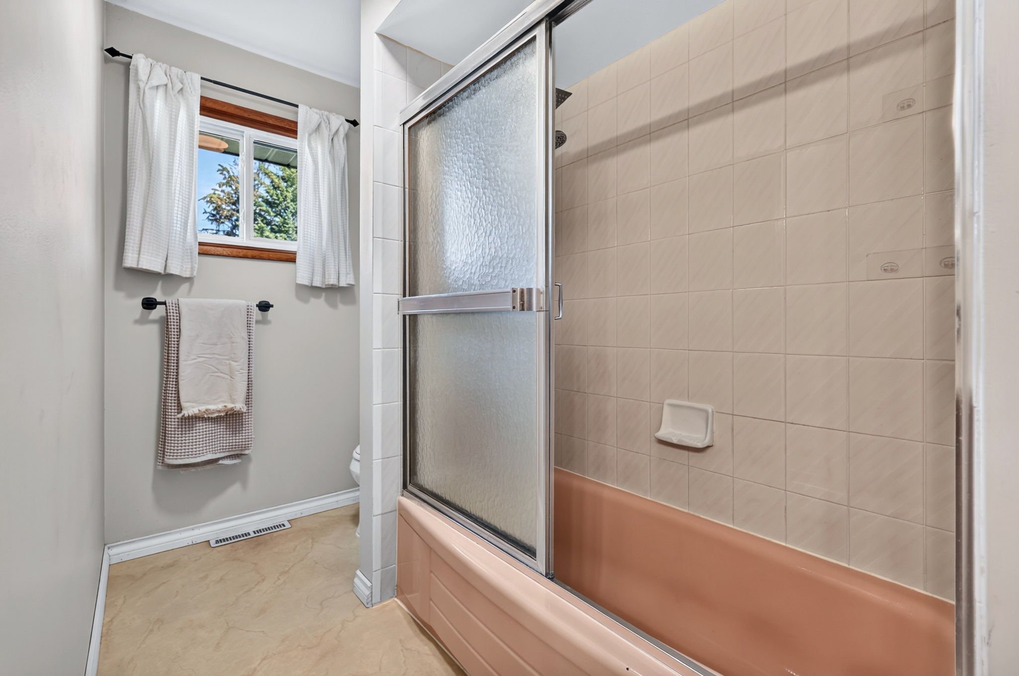 Bathroom with beige tiled shower stall, window with white curtains, towel rack with towels, and a small wall vent.