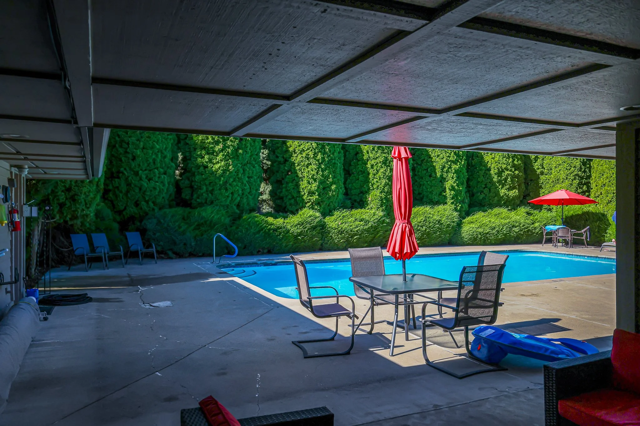 View of a backyard swimming pool area with outdoor furniture, umbrellas, and lush green trees in the background, partially shaded by a covered patio ceiling.