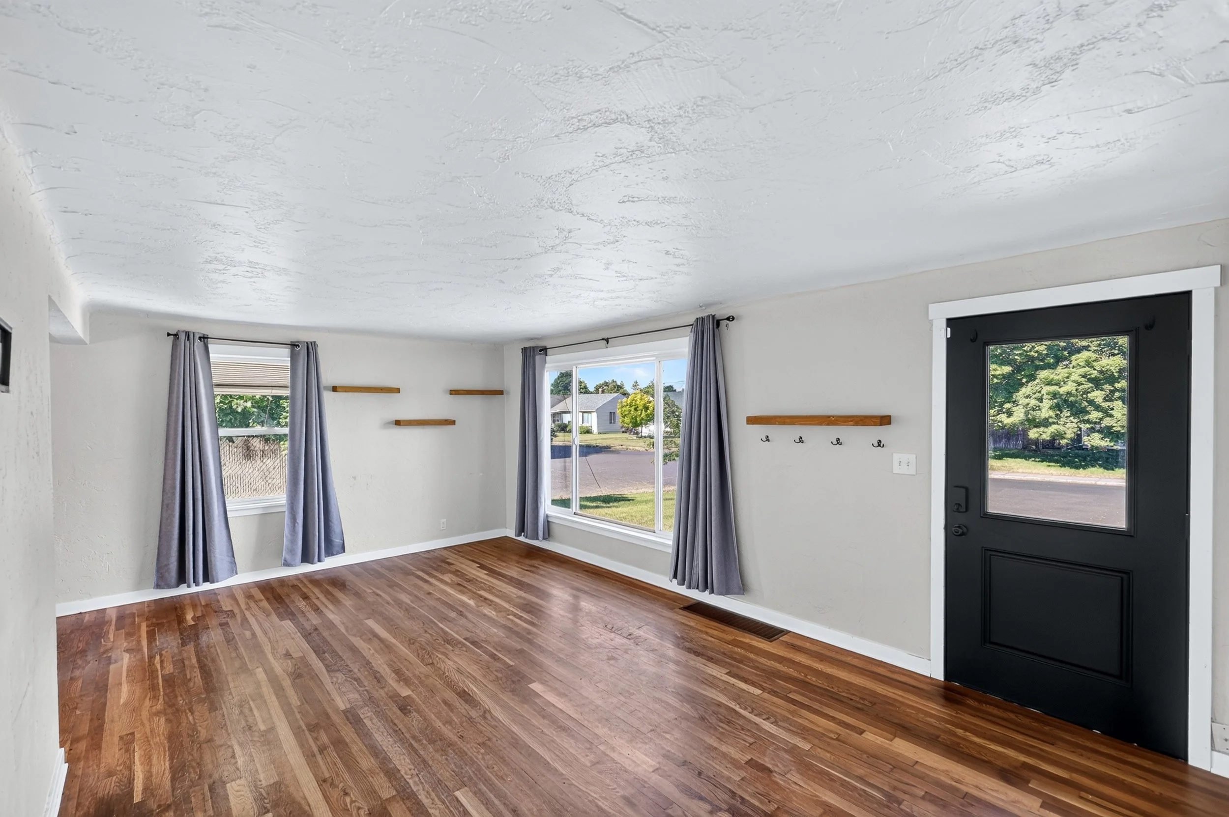 Empty living room with hardwood floor, white walls, three large windows with gray curtains, and a black front door with glass panel.