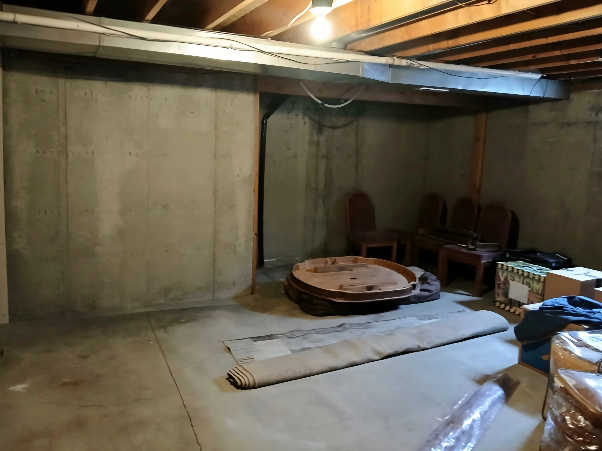 A basement with concrete walls and floor, with chairs, boxes, and miscellaneous items scattered around. A single lightbulb hanging from the ceiling illuminates the room.
