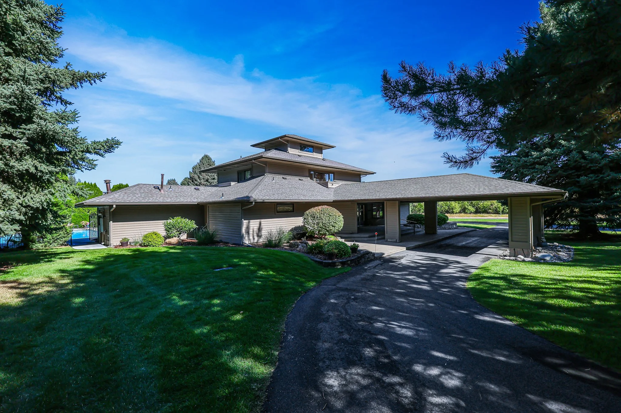 A large house with a multi-tiered roof, surrounded by lush green lawn, trees, and shrubs, under a bright blue sky with some clouds.