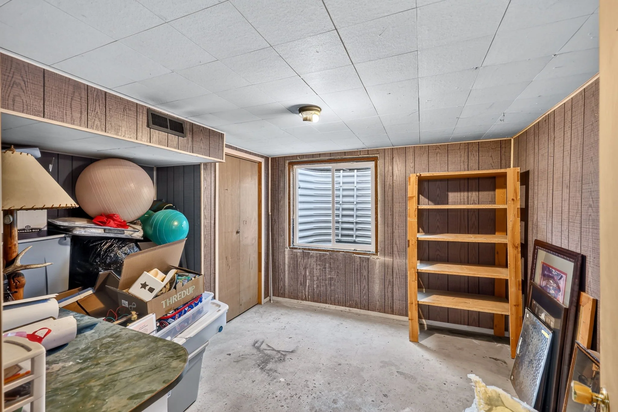 A cluttered basement room with wood paneled walls, a window, and various storage items including exercise balls, a lamp, and a bookshelf.