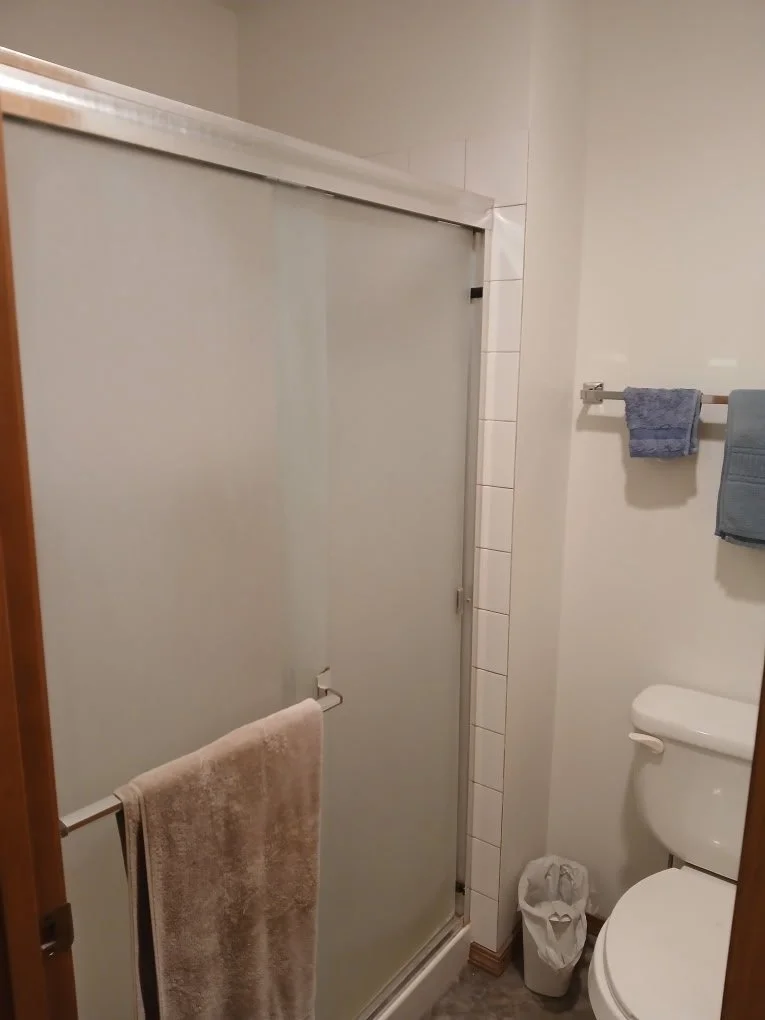 Bathroom with a glass shower door, beige towel hanging on a towel bar, blue towel on a wall-mounted rack, toilet with a small trash can beside it, and tiled walls.