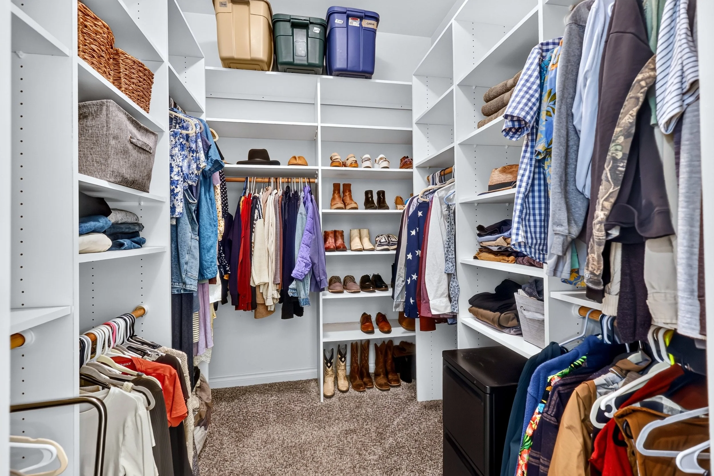 A walk-in closet with white shelves filled with folded clothes, shoes, and storage bins, as well as hanging shirts, dresses, and pants.