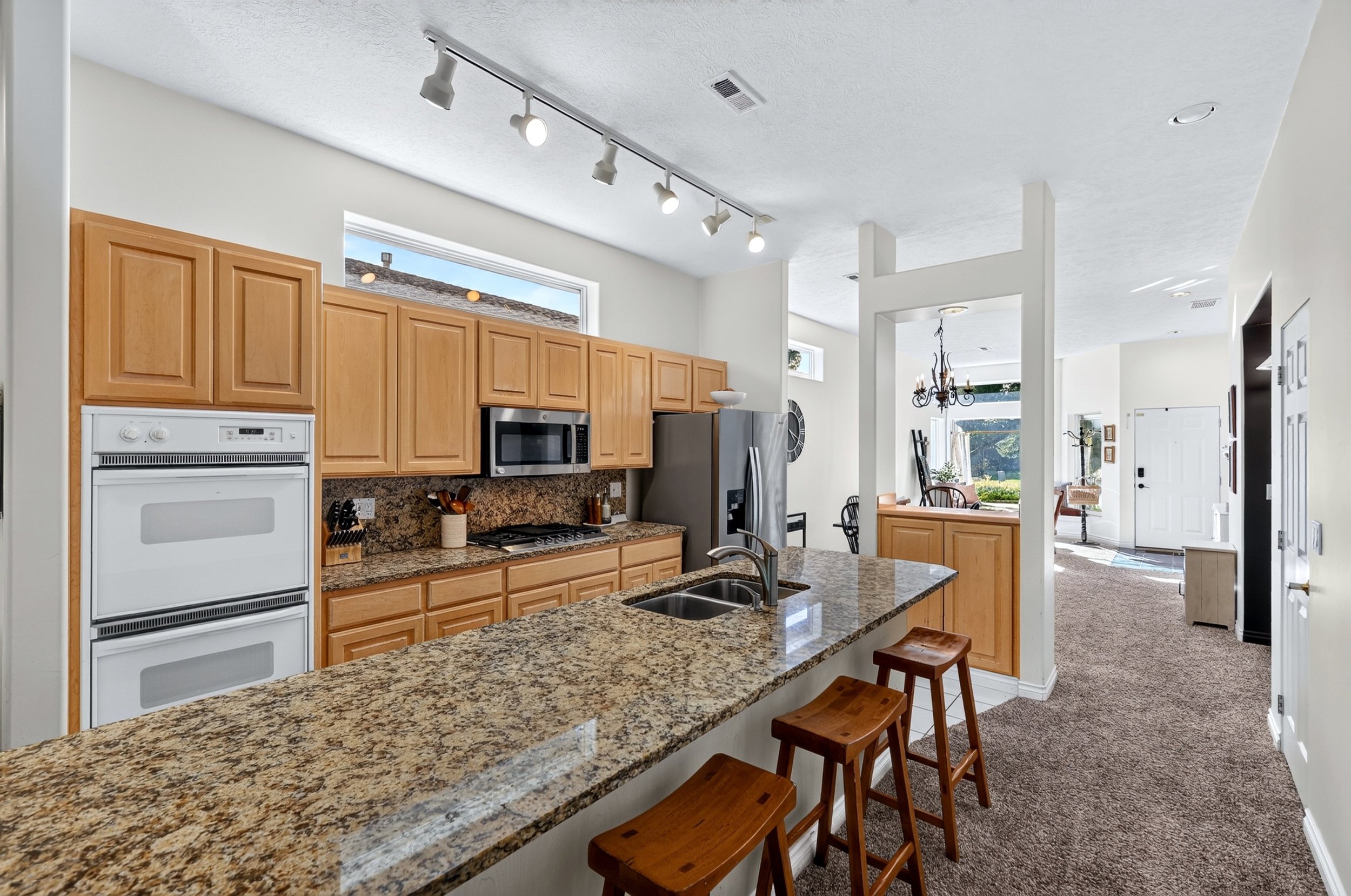 Kitchen with granite countertops, wooden cabinets, stainless steel appliances, including a refrigerator, microwave, and oven, and a breakfast bar with stools. Brightly lit with natural light and ceiling track lighting, opening into a dining and livin