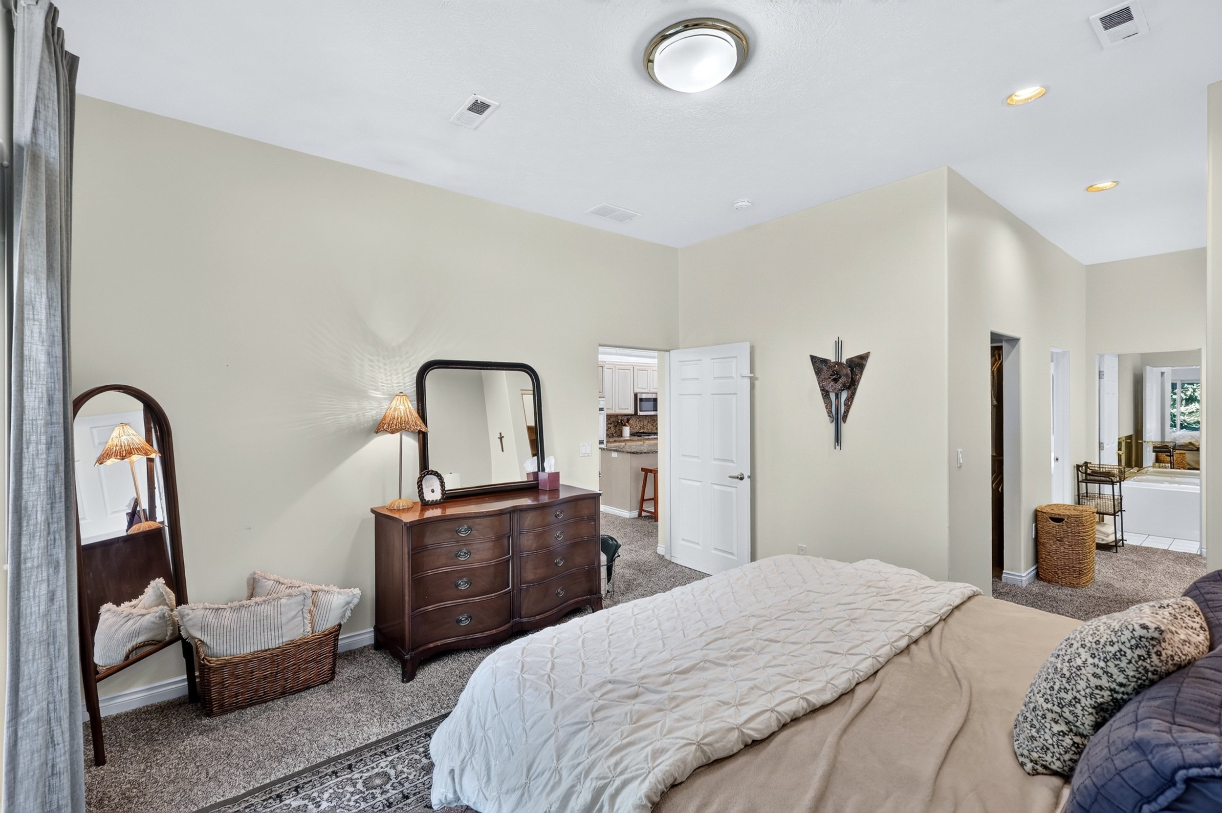 A bedroom with a bed, a wooden dresser with a mirror, and a basket on the floor. The room is decorated with two lamps, a cross, and a wall sculpture. The walls are beige, and there are multiple doors leading to a kitchen and an adjacent room.