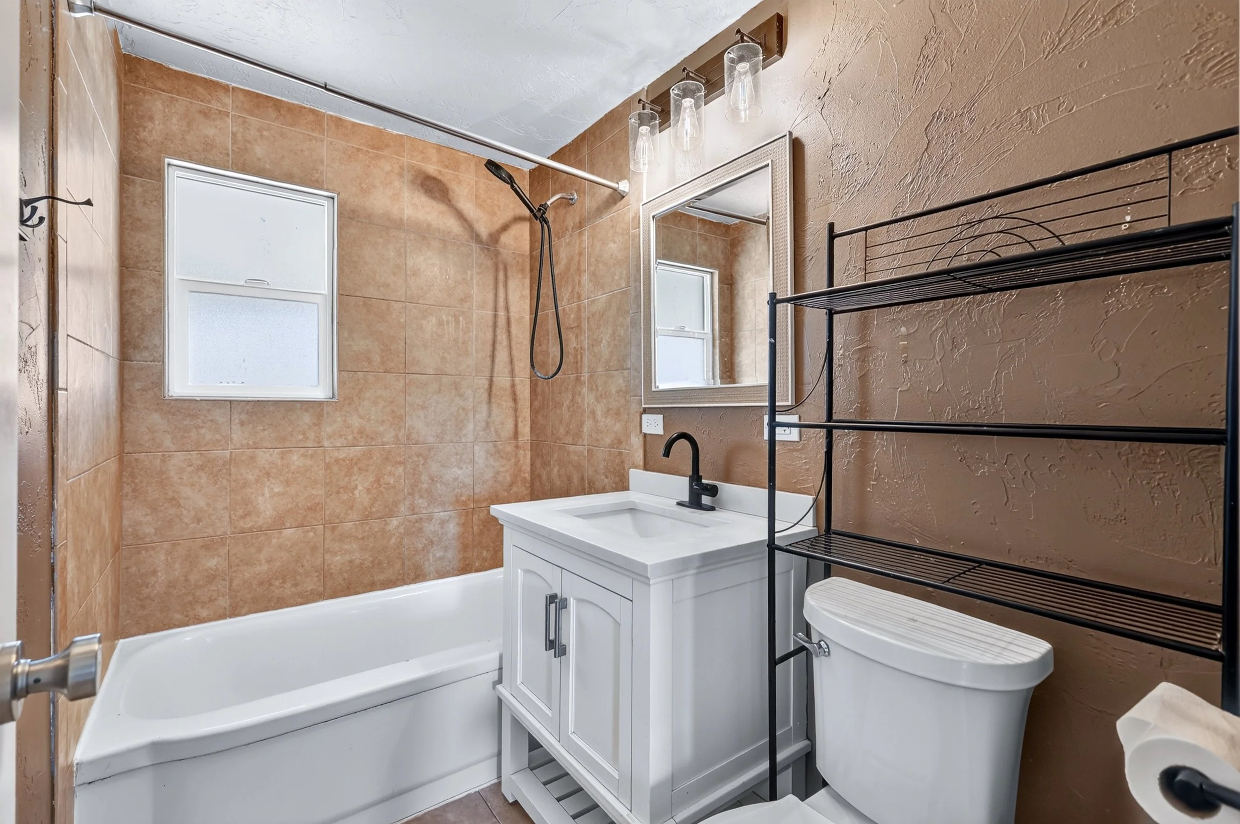 A small bathroom with tan tiled walls, a clawfoot bathtub, a white vanity with a sink, a mirror, frosted window, black metal shelving, and a textured brown wall.