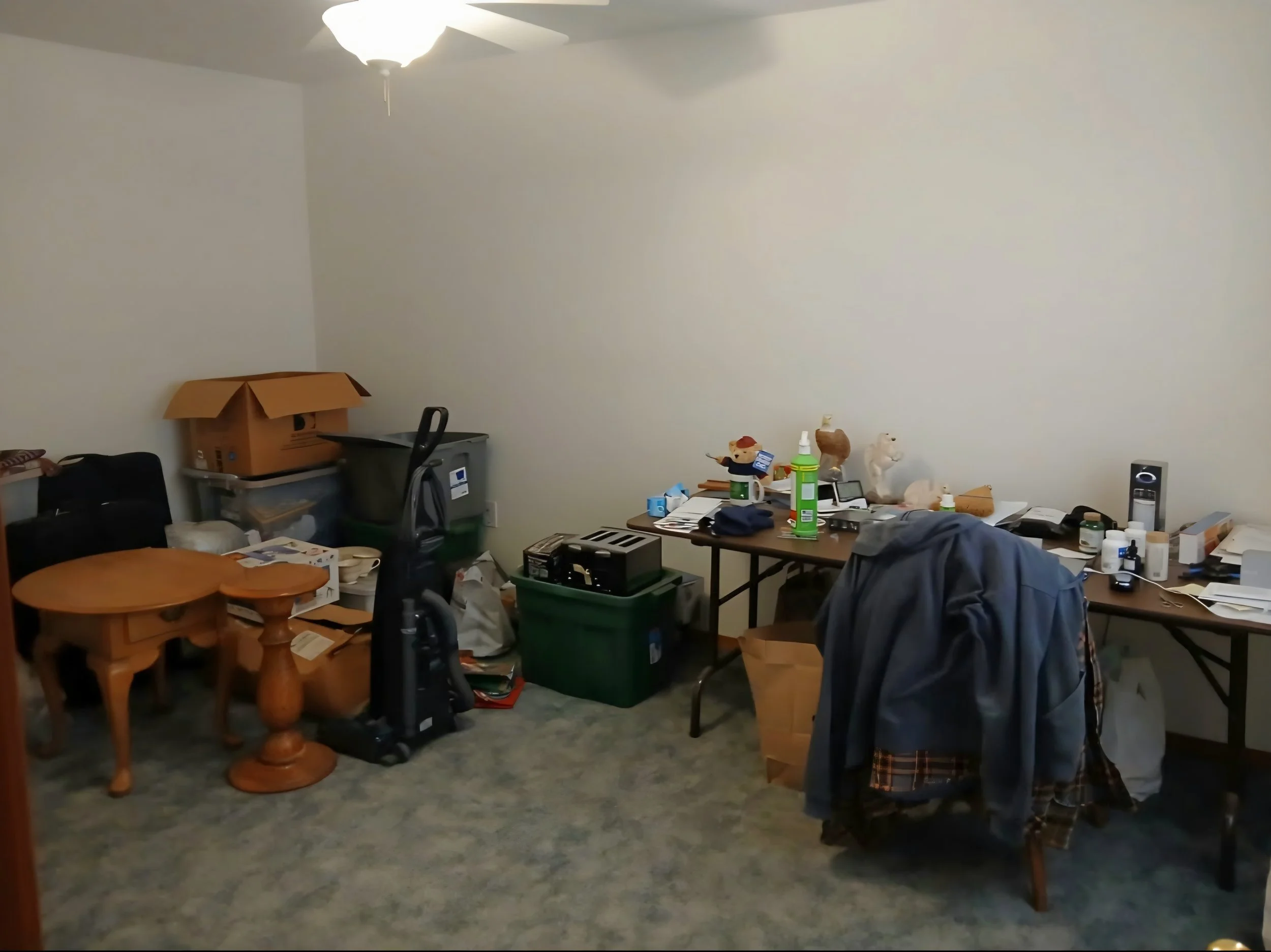 Room filled with clutter, including a table with various objects, chairs, boxes, and stuffed animals, under a ceiling fan with a light.