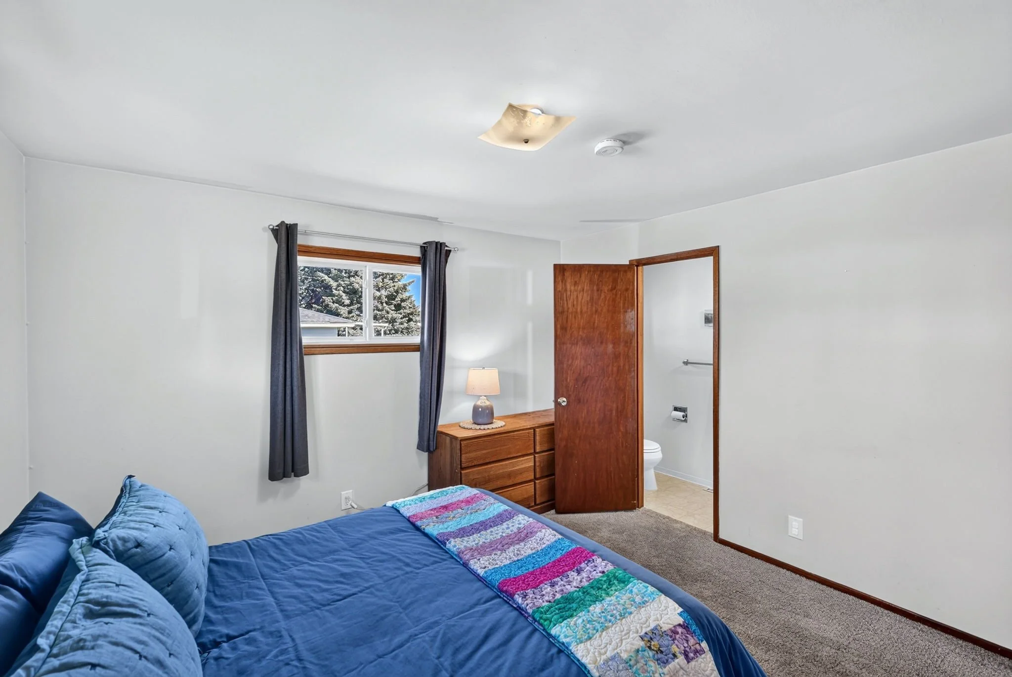 A bedroom with a blue bed and a colorful quilt, a wooden dresser near a window with dark curtains, and a ceiling light fixture. An open door reveals a bathroom with a toilet and sink.