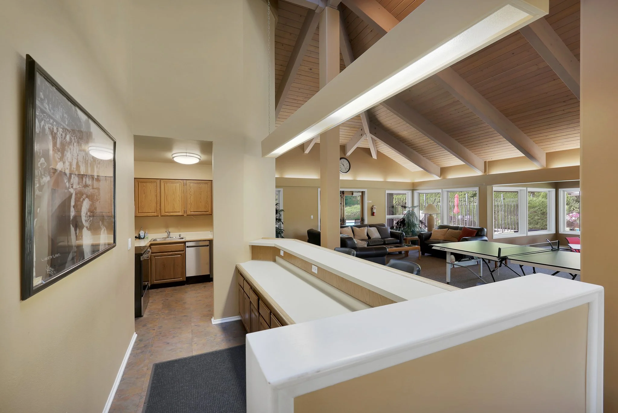 View from a corner of a room showing a kitchen area with wooden cabinets, a sink, and a countertop, overlooking a living room with multiple large windows, leather sofas, and a ping pong table. The ceiling has wooden beams and skylights, and there is 