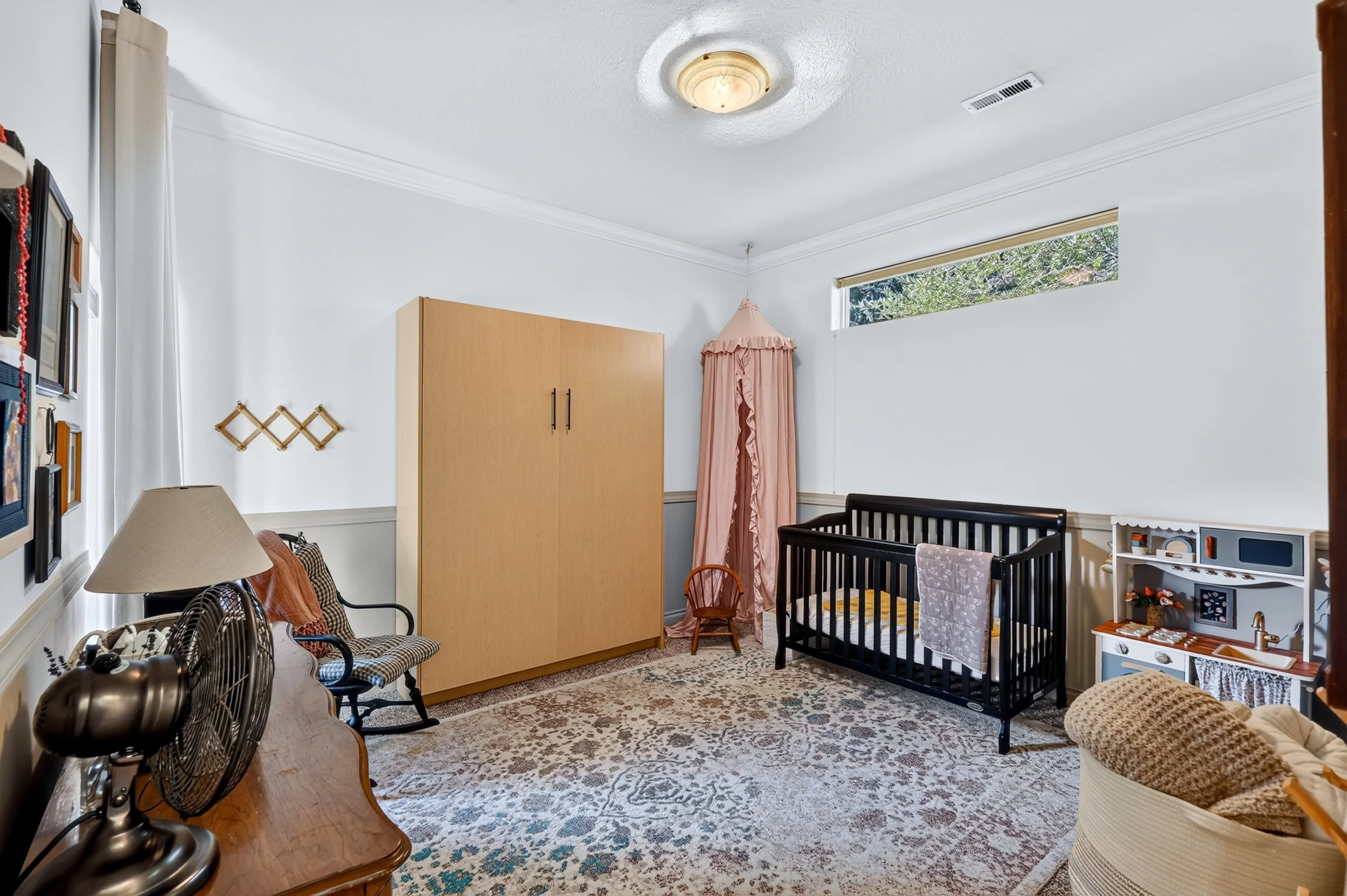 Living room with white walls, a patterned rug, a black crib, a small wooden chair, a pink canopy, a large wooden wardrobe, a patterned armchair, a lamp, and various decorations on the wall.