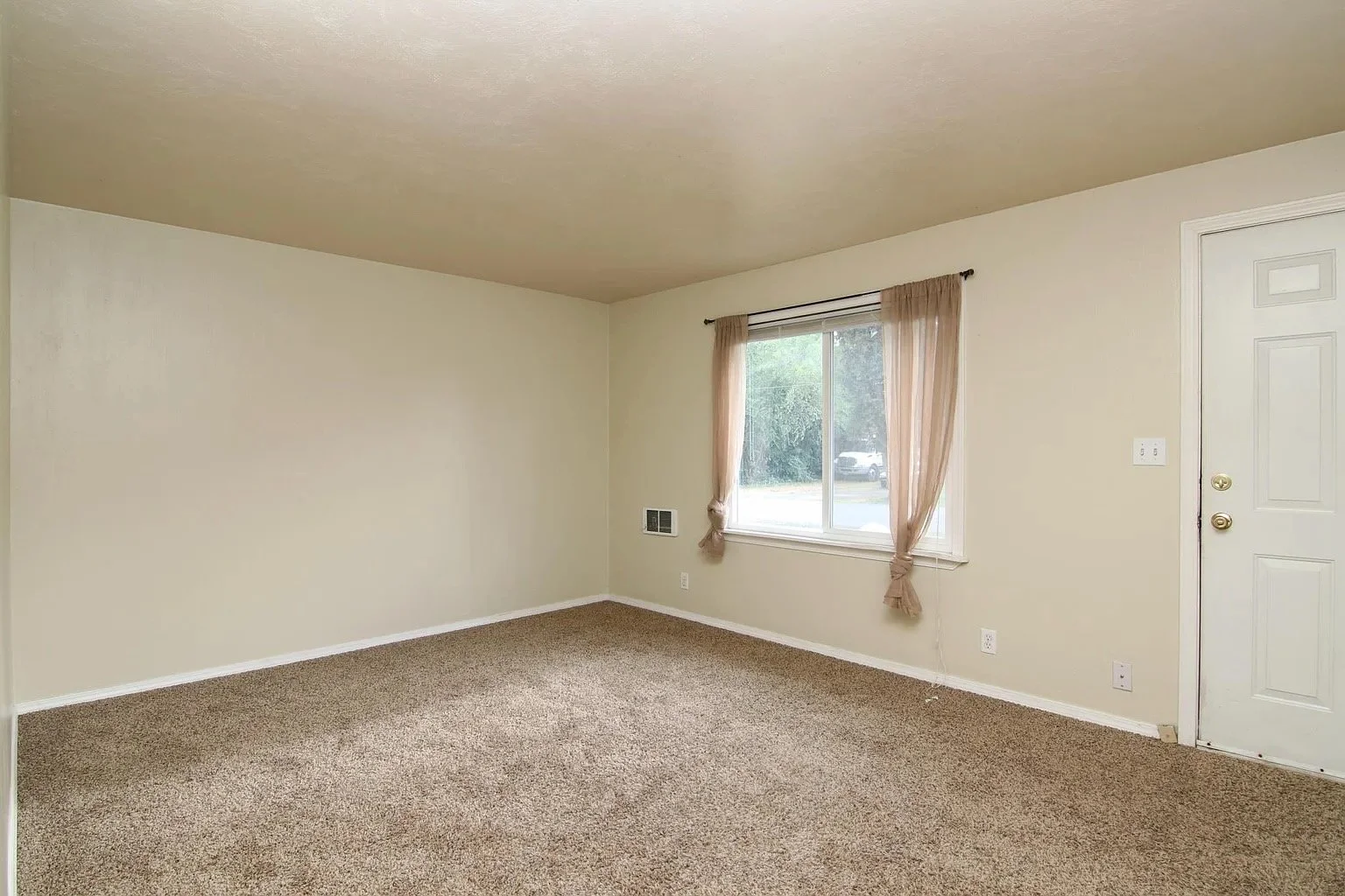 Empty living room with beige walls, brown carpeted floor, a window with beige curtains, and a white door.