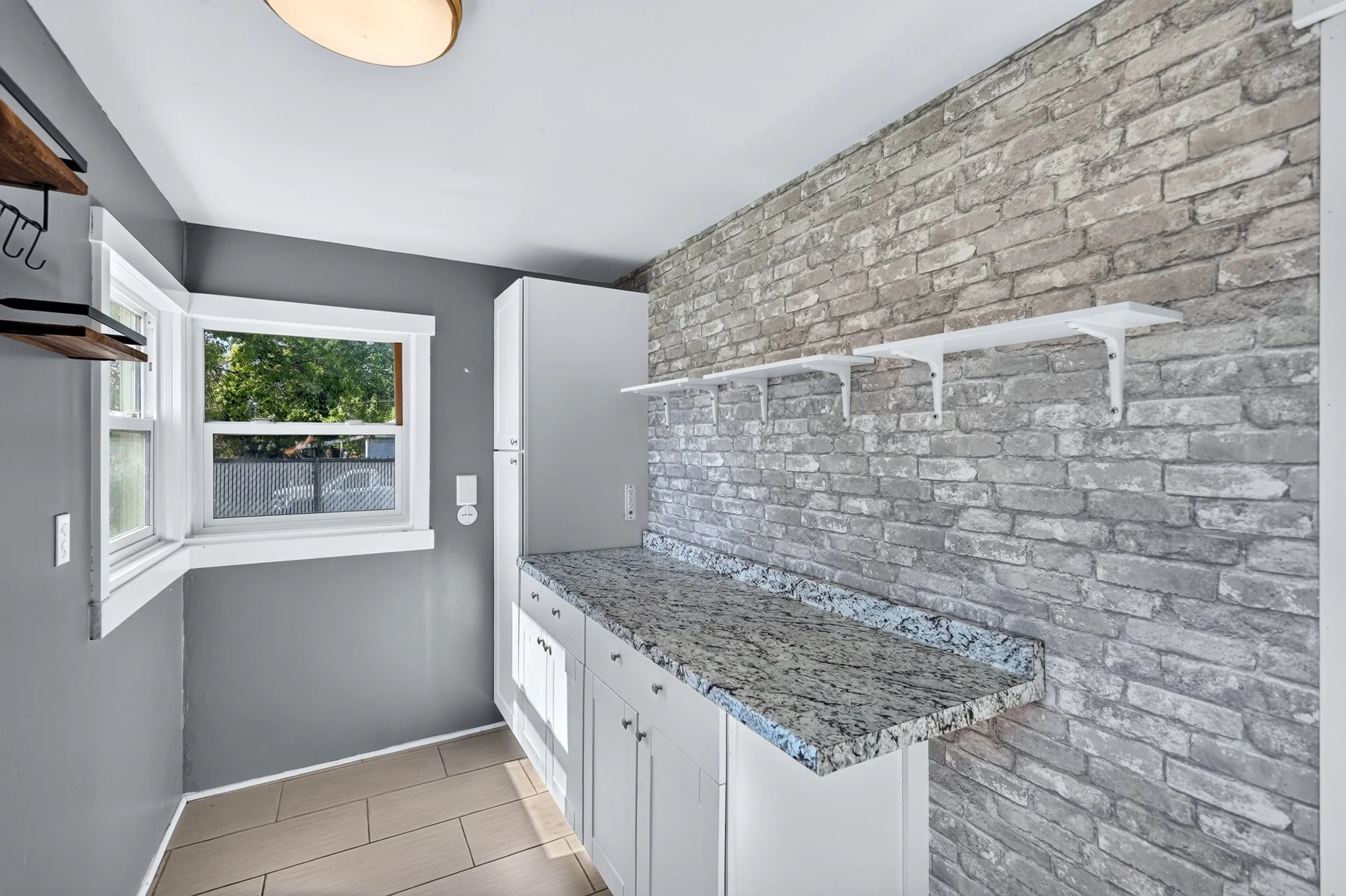 Modern kitchen with gray walls, white cabinetry, granite countertops, a brick accent wall, and a window overlooking trees.