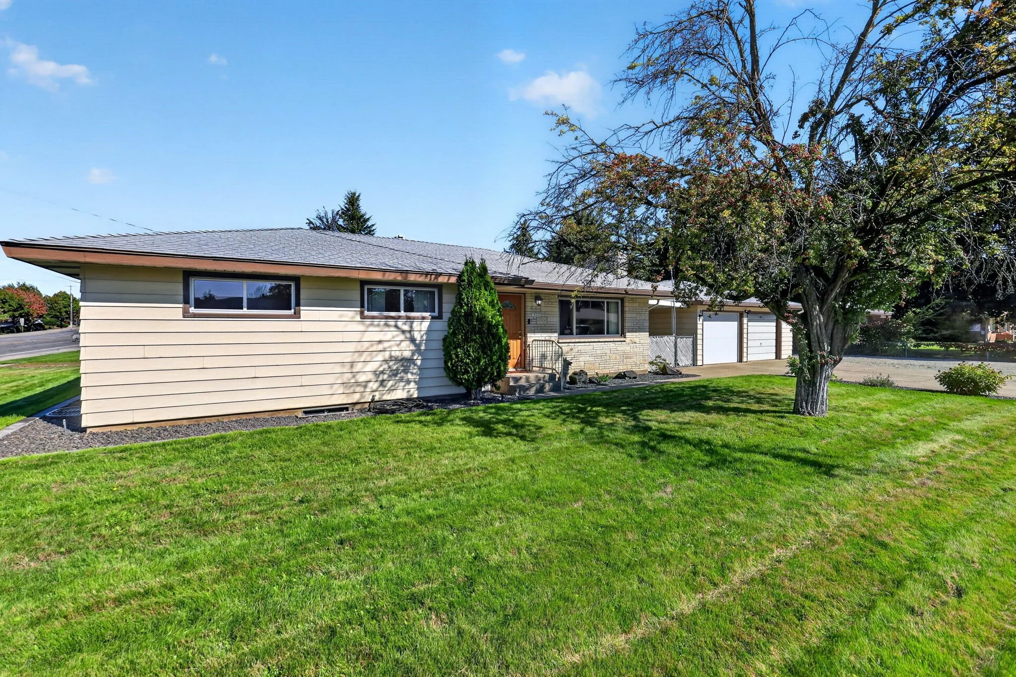 A suburban house with a brick and siding exterior, two horizontal windows, and a brown front door. There is a large tree in the front yard with green grass and some orange and green leaves. The sky is blue with a few small clouds.