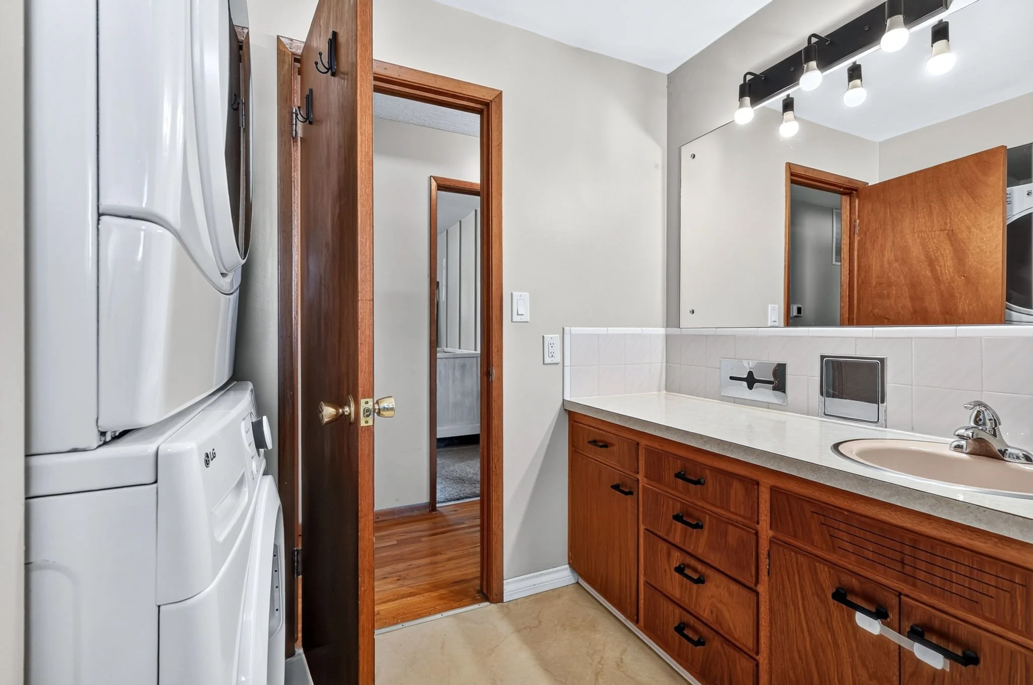 A laundry area with stacked washer and dryer on the left, wooden cabinets and a sink on the right, and above the sink a mirror with a modern light fixture. Open doorways lead to a hallway and another room.