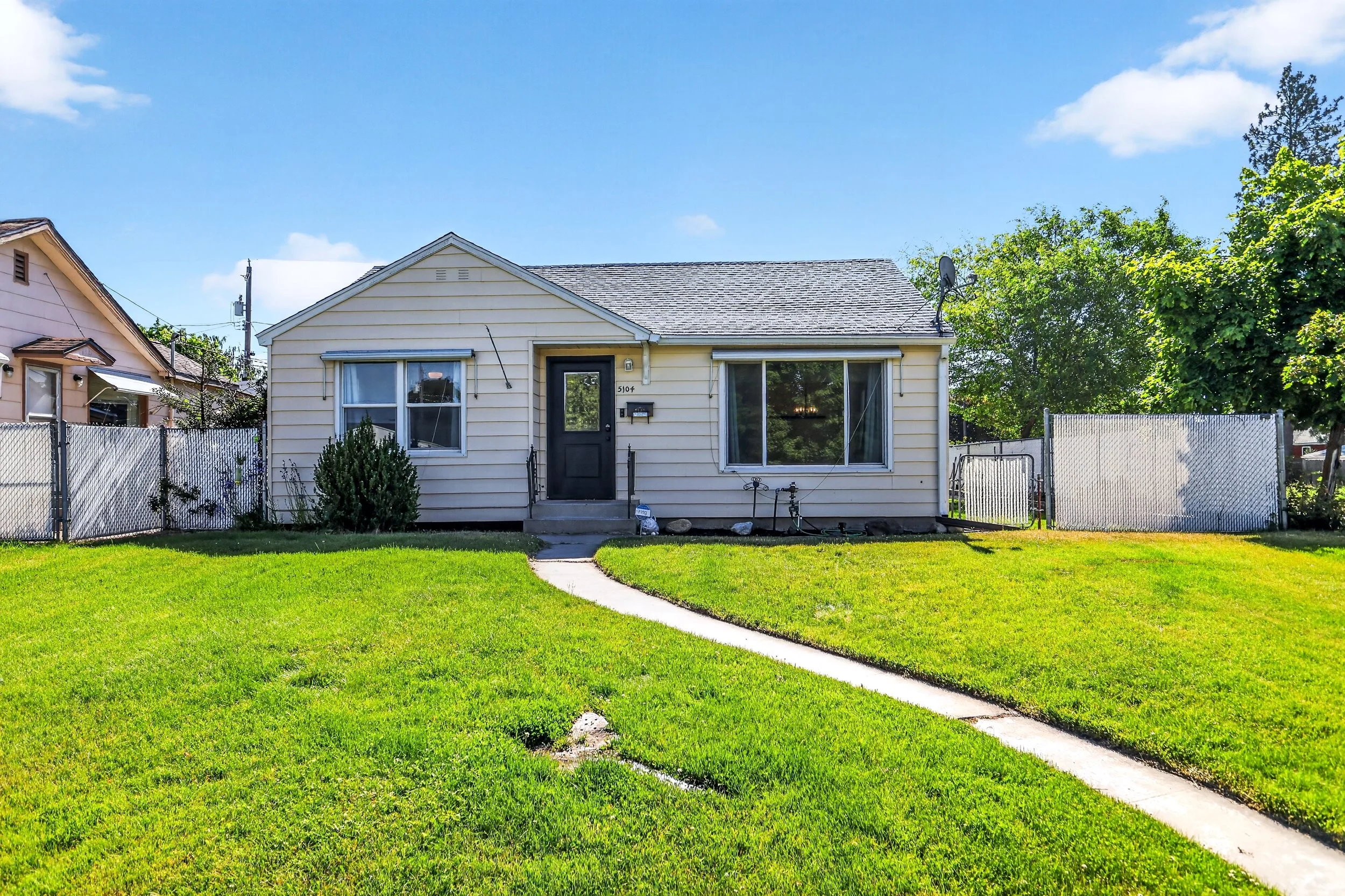A small, yellow, single-story house with white trim, sitting on a green lawn with a concrete walkway leading to the front door. The house has two windows, a front porch with steps, and a satellite dish on the roof. There are trees and neighboring hou