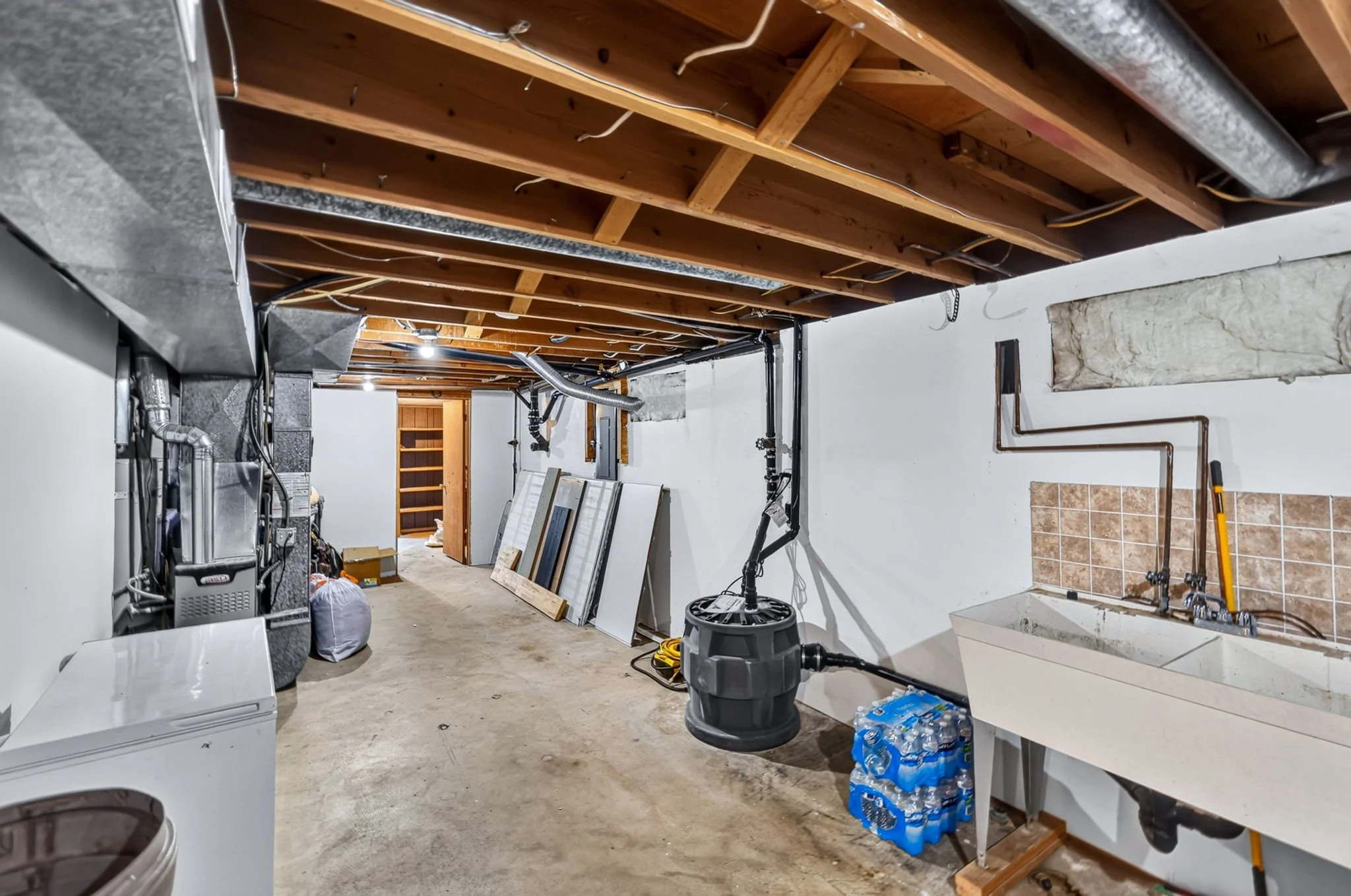 Unfinished basement with exposed wooden ceiling beams, concrete floor, and various tools and supplies like bottled water, a vacuum, and old tiles.