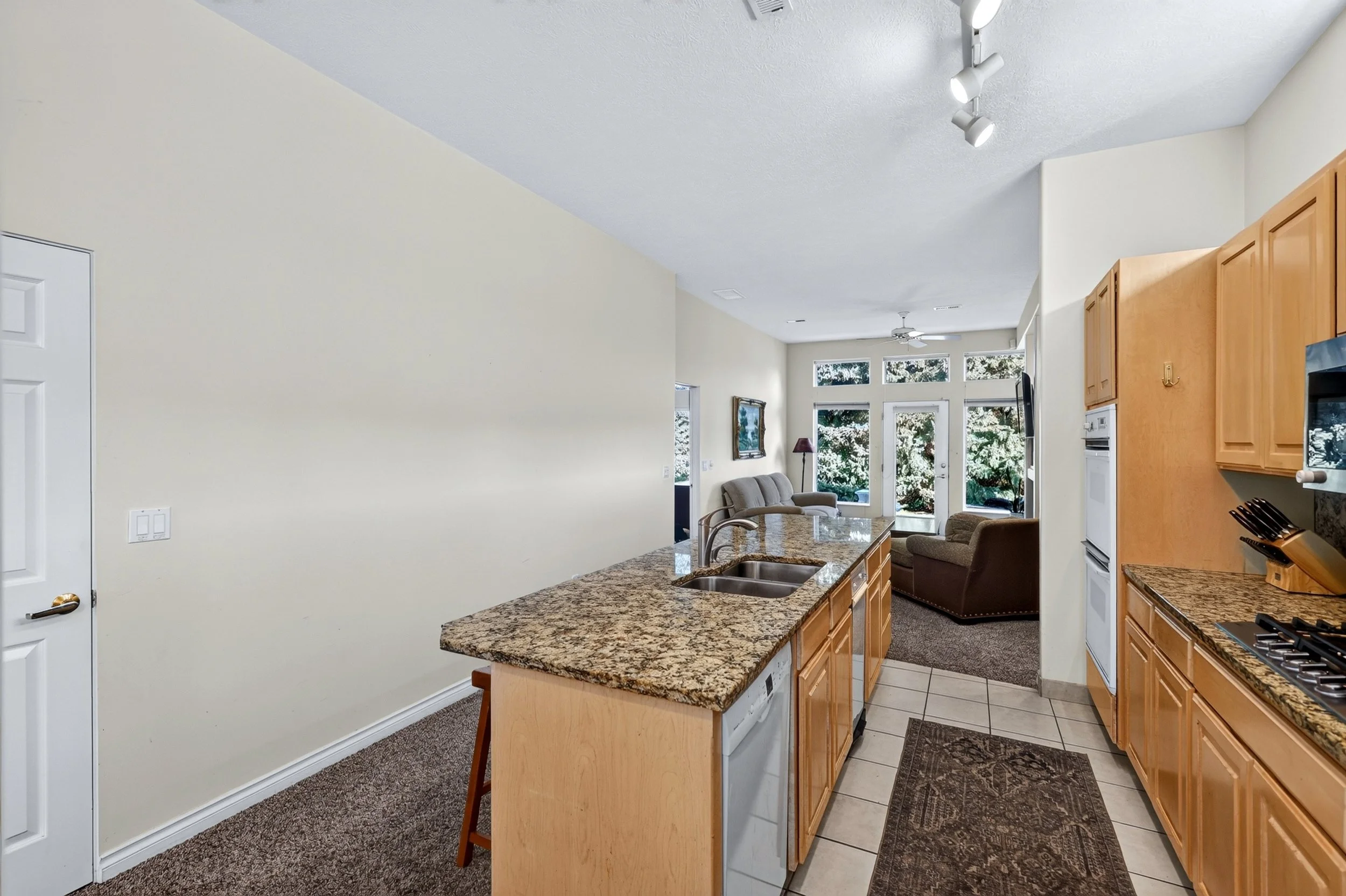 Open floor plan kitchen and living room area with granite countertops, wooden cabinets, large windows, ceiling fan, and furniture.