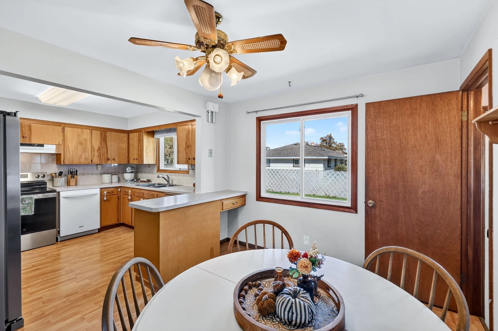 Kitchen with wooden cabinets, a stainless steel refrigerator, a white stove, and a window showing an outside yard. A round dining table with a floral centerpiece is in the foreground.